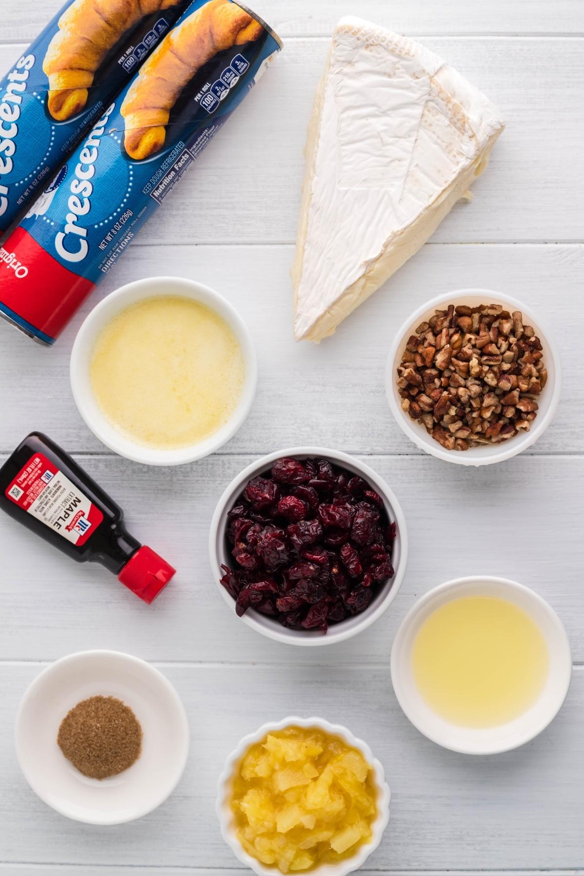 Crescent rolls, brie, pecans, dried cranberries, butter, vanilla, pineapple juice, and spices come together to create a festive cranberry brie wreath on a white table.