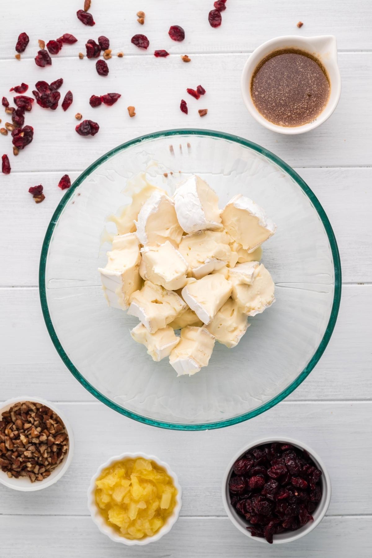 Glass bowl with cubed brie cheese, surrounded by small bowls of pecans, pineapple, cranberries, and sauce—perfect ingredients for a festive Pineapple Cranberry Brie Wreath or a tasty crescent wreath appetizer.