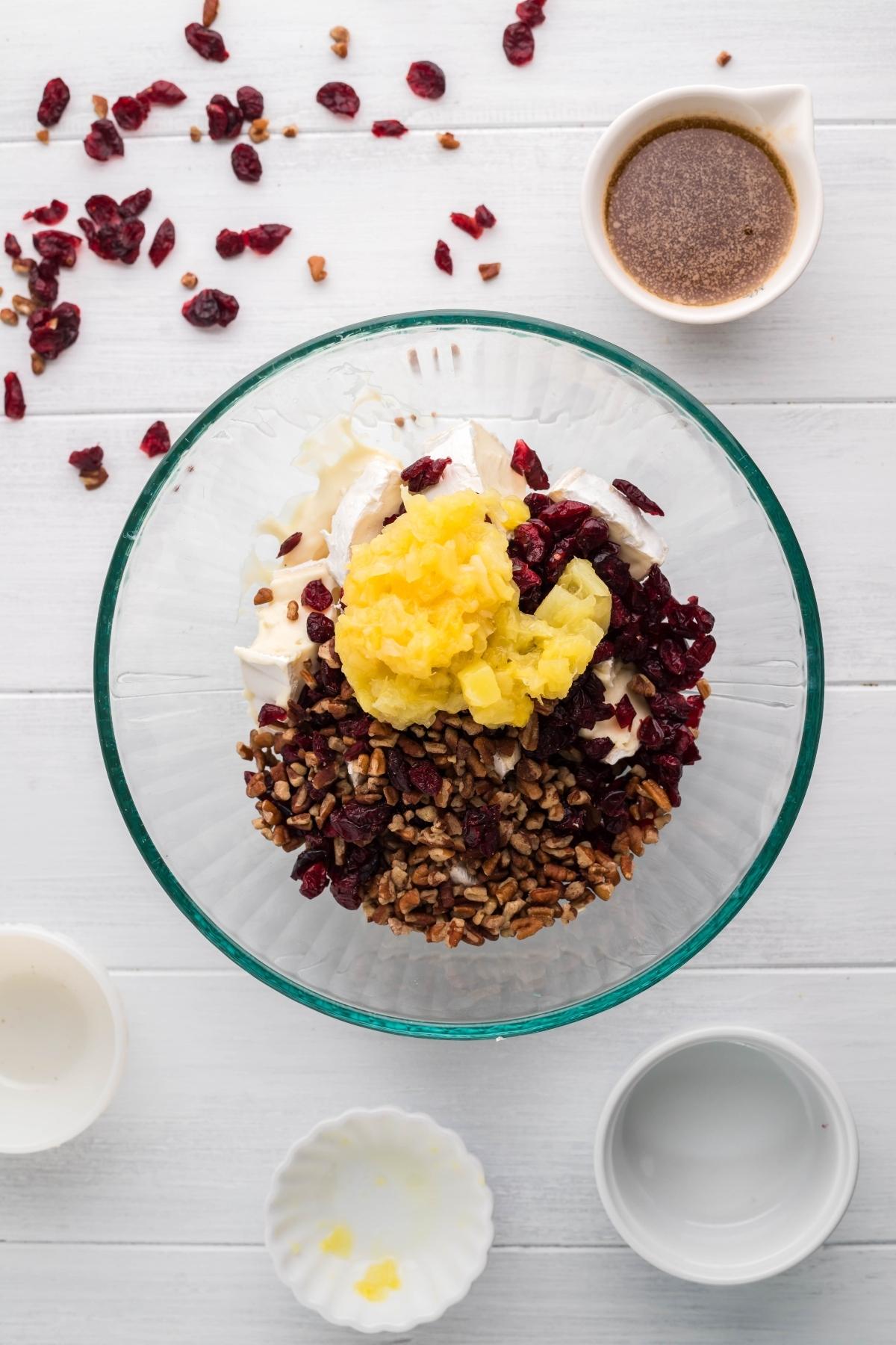Glass bowl with cream cheese, pineapple, pecans, and cranberries, surrounded by small bowls on a white table—perfect as a spread for your crescent wreath appetizer or Pineapple Cranberry Brie Wreath.
