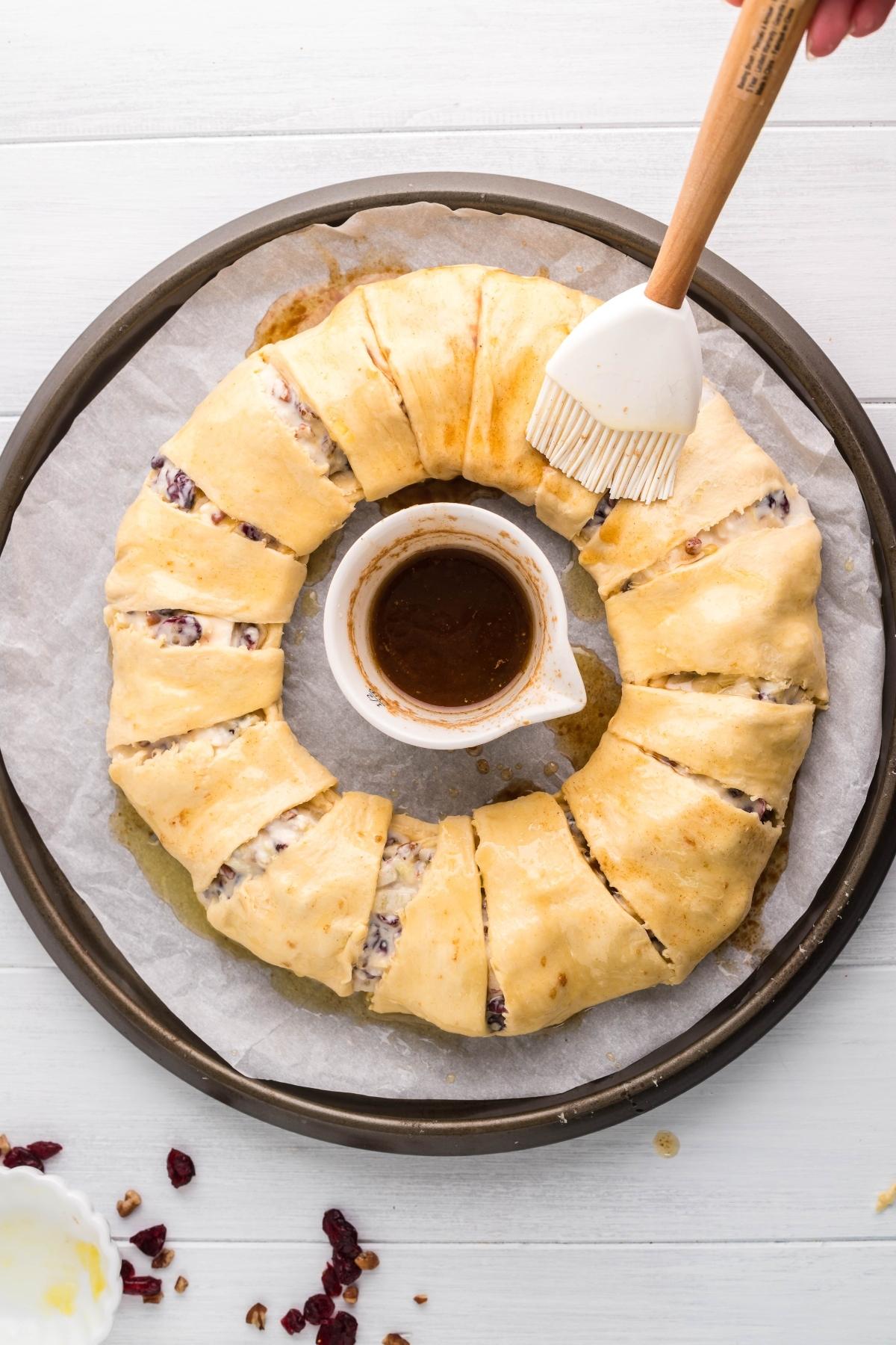 A hand brushes glaze onto a sliced Pineapple Cranberry Brie Wreath on a round baking tray with a bowl of glaze in the center.