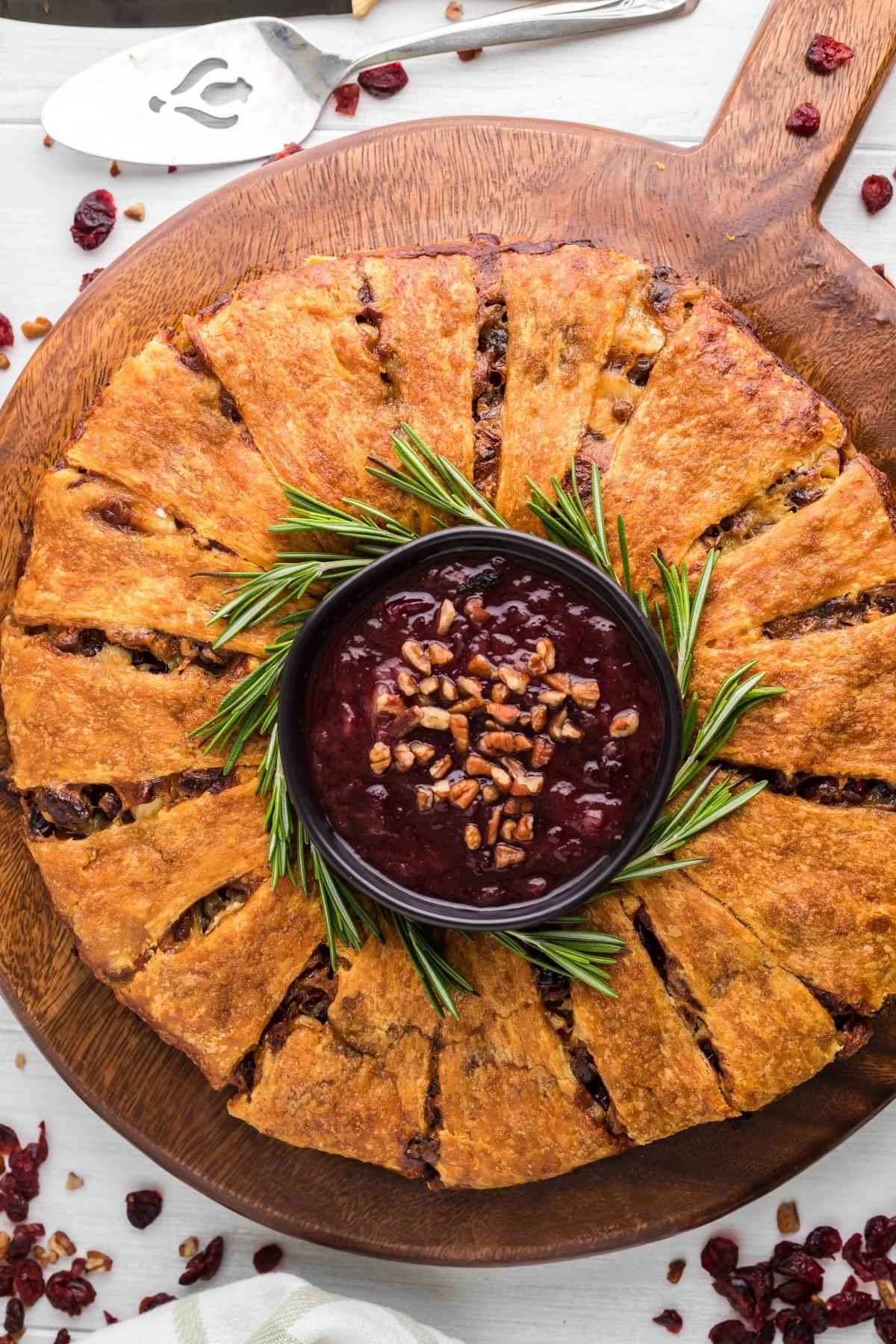 Round baked bread ring with dipping sauce in the center, garnished with rosemary on a wooden board—this festive crescent wreath appetizer is perfect for any gathering.