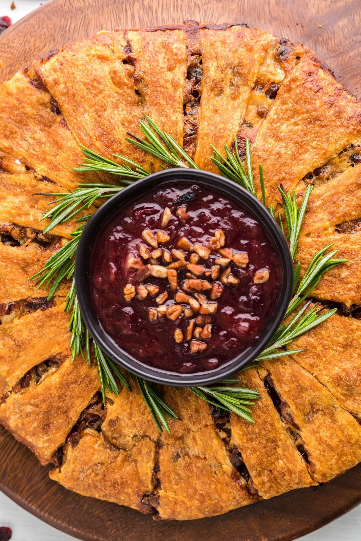 Golden baked cranberry brie wreath, shaped like a festive wreath, served with berry sauce, pecans, and rosemary for a delightful touch.