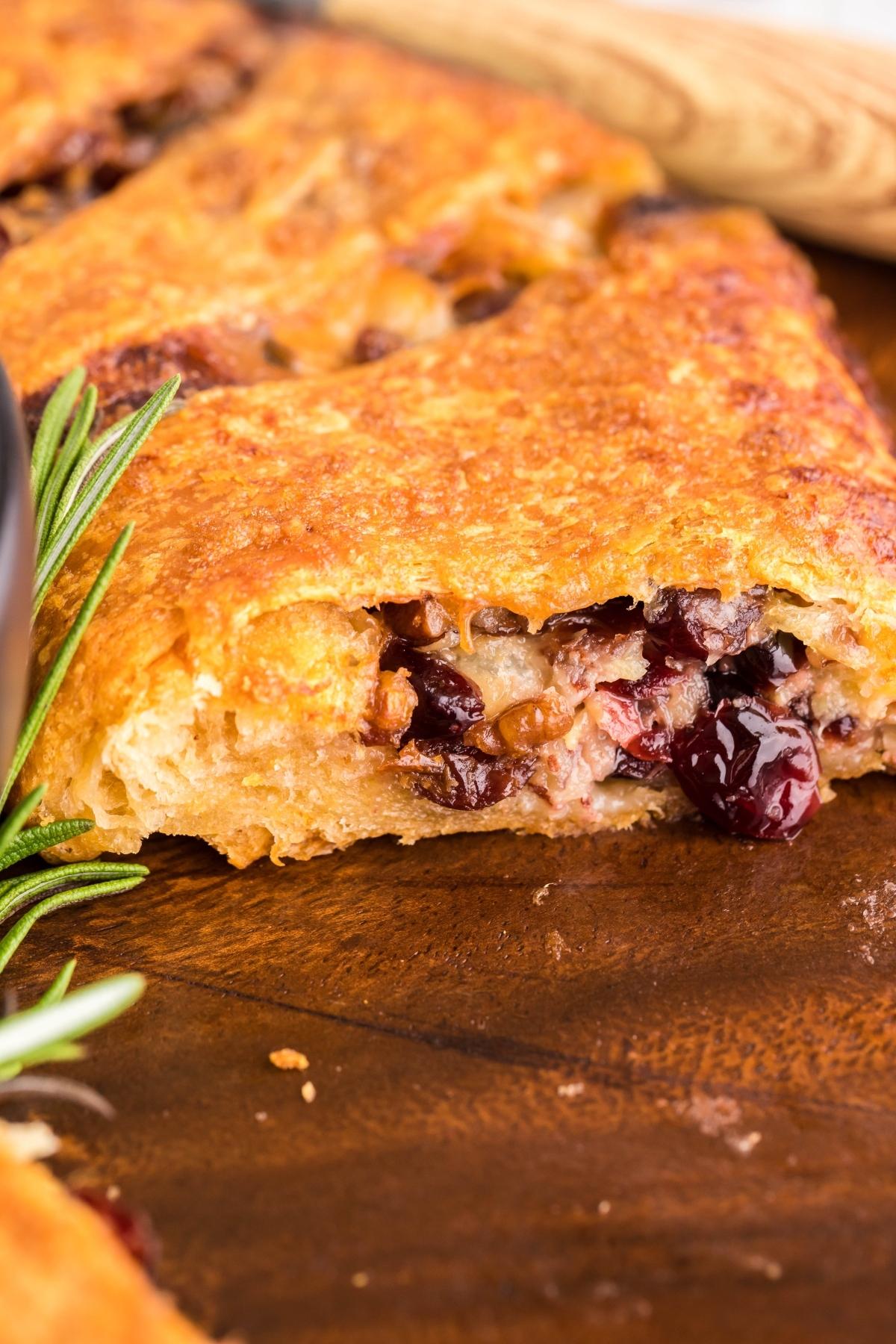 A close-up of a flaky pastry inspired by a crescent wreath appetizer, filled with cranberries and nuts on a wooden surface, garnished with fresh rosemary.