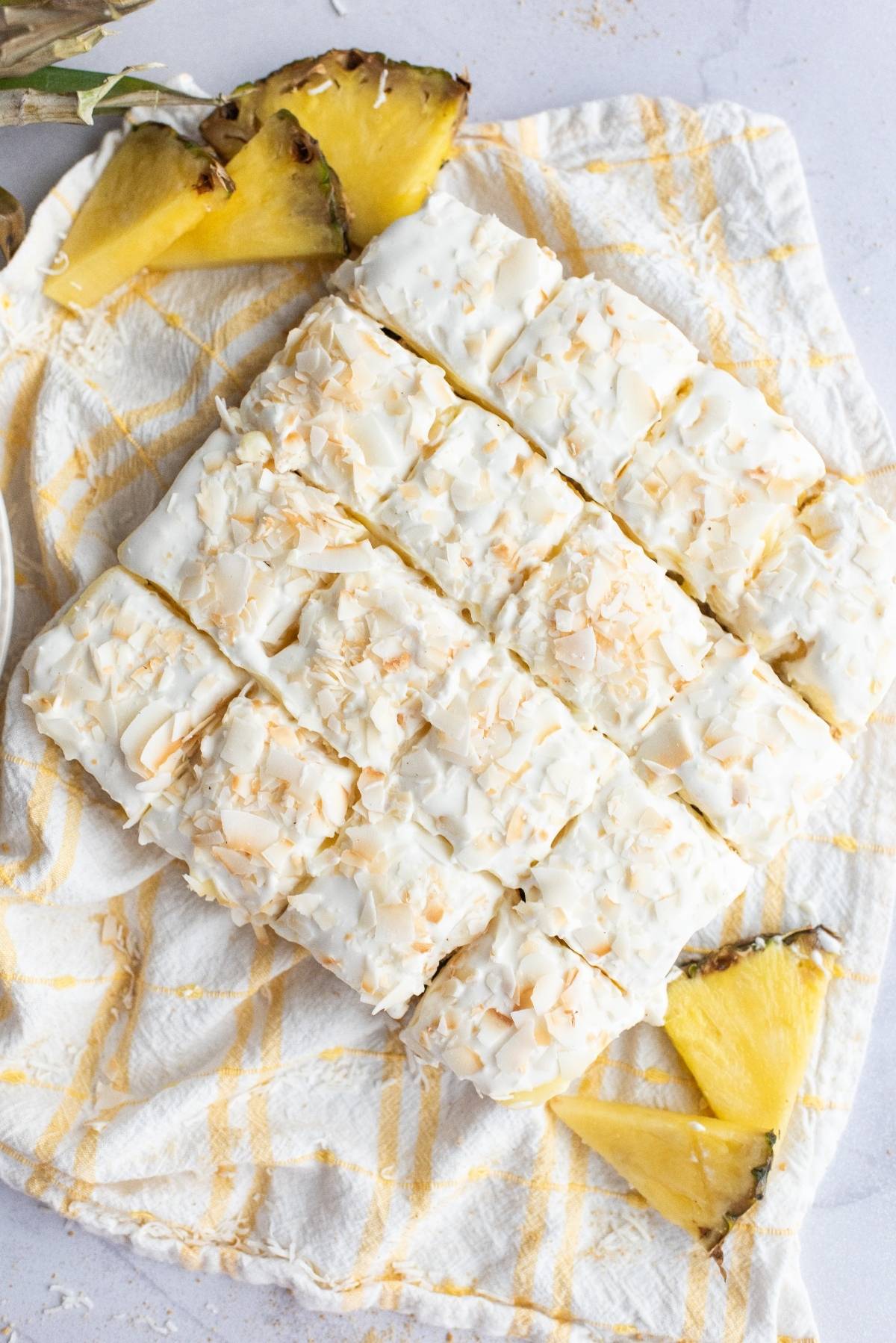 Nine frosted Hawaiian pineapple and coconut cake bars, topped with coconut flakes, arranged on a cloth with pineapple slices nearby.