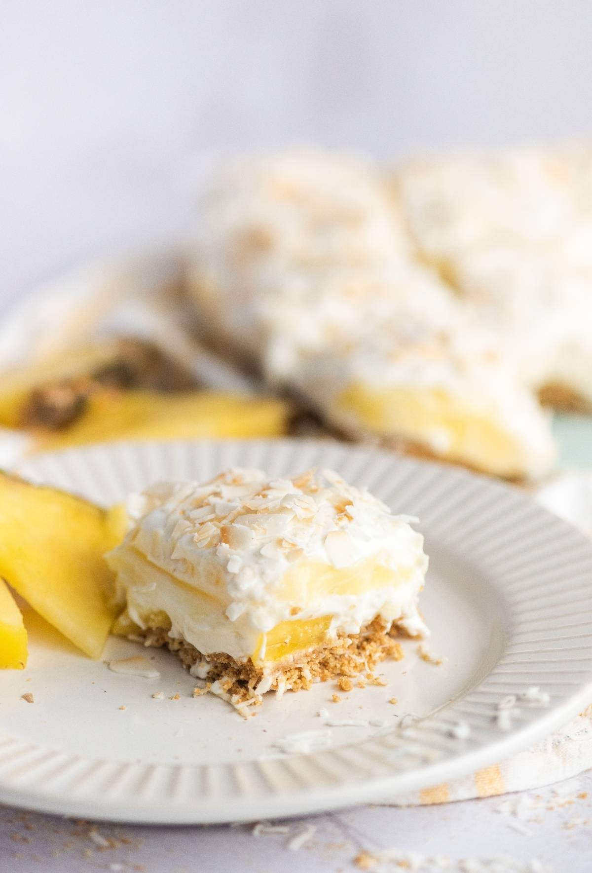 A slice of creamy summer dessert—Hawaiian pineapple and coconut cake—topped with whipped cream and toasted coconut, served alongside pineapple slices on a white plate.