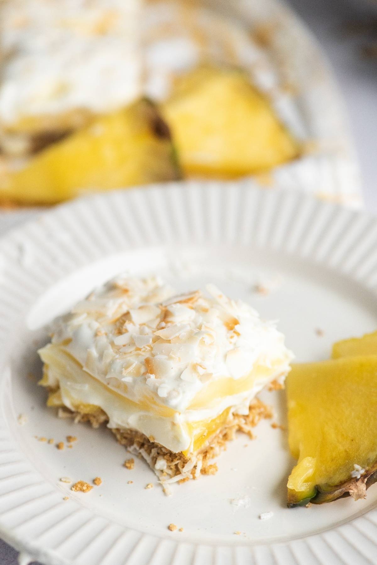 A slice of creamy summer dessert topped with whipped cream and toasted coconut on a white plate with pineapple wedges—a perfect no bake cake recipe for a summer BBQ.