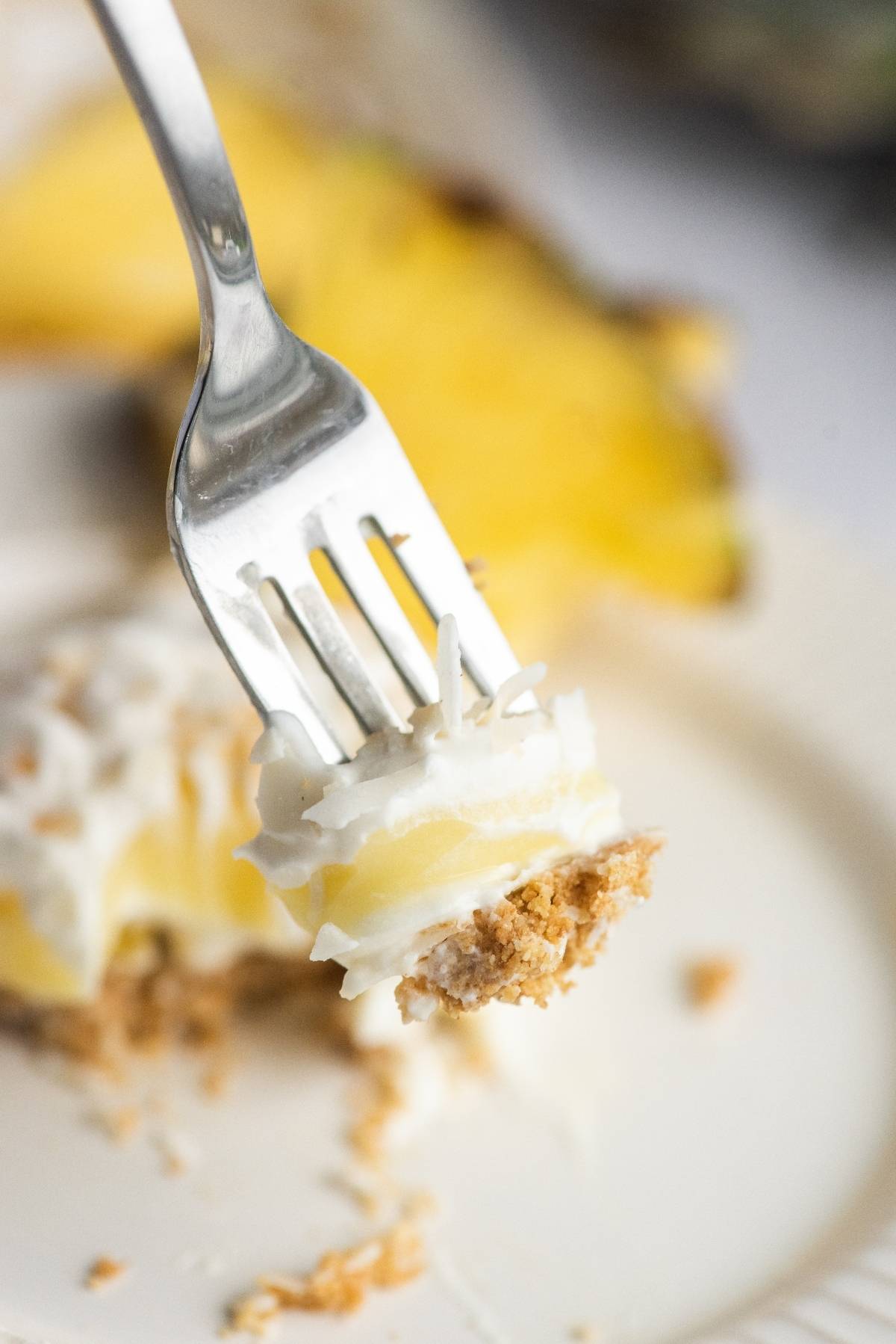 A fork holds a bite of creamy summer dessert with crust and pineapple, above a white plate—perfect as a no bake cake recipe for your next summer BBQ.