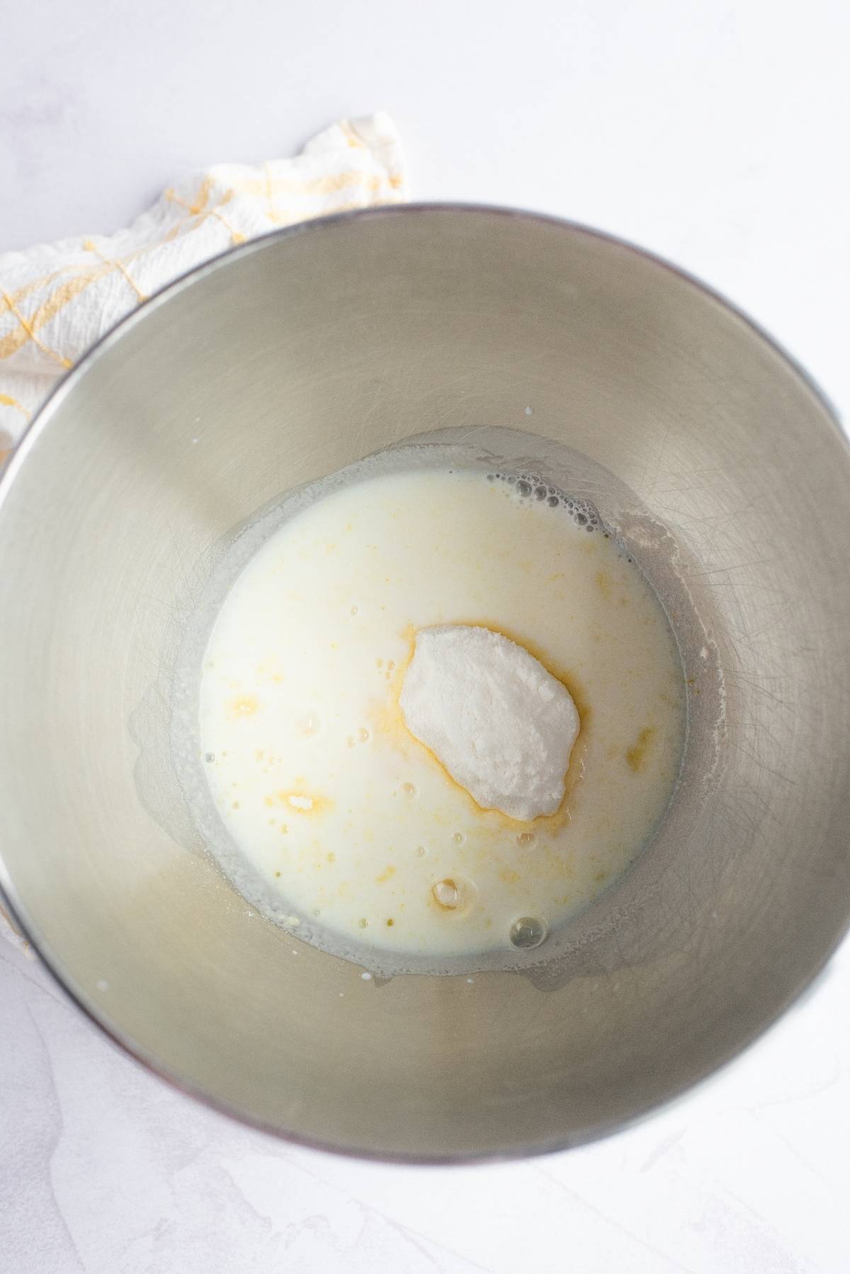 A metal mixing bowl with milk and a mound of white powder on a light surface, next to a towel—perfect for whipping up a creamy summer dessert like pineapple lush, an ideal dessert for a summer BBQ.