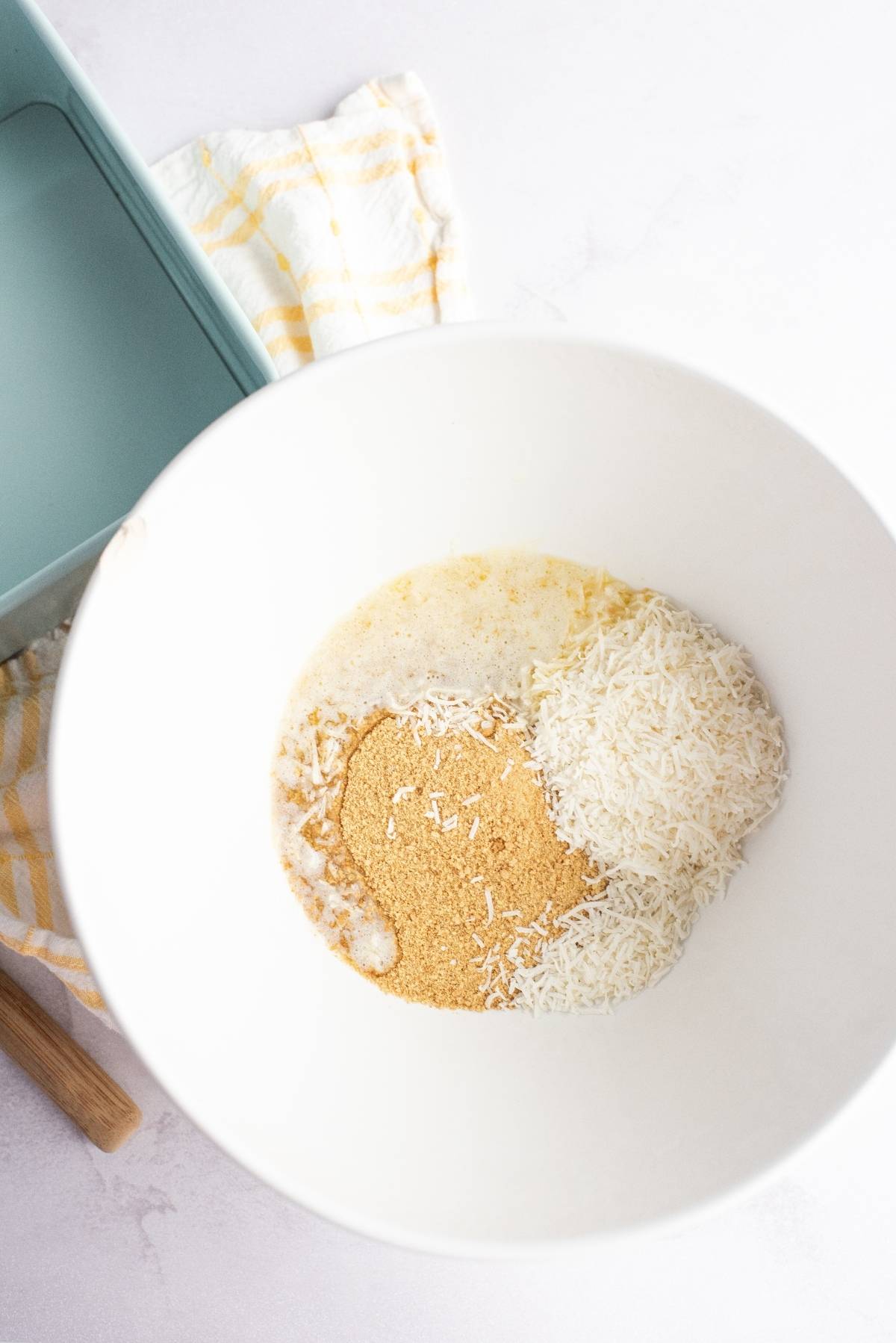 White bowl with shredded coconut, graham cracker crumbs, and melted butter next to a blue baking dish—perfect ingredients for a no bake cake recipe like pineapple lush, an ideal dessert for a summer BBQ.