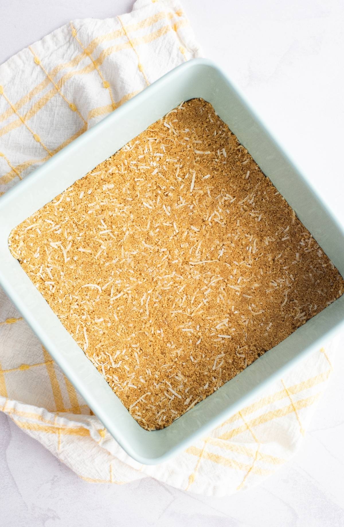A square baking dish with a crumbly, golden crust—this pineapple lush dessert sits atop a white and yellow checkered cloth, ready to impress lovers of Hawaiian pineapple and coconut cake.