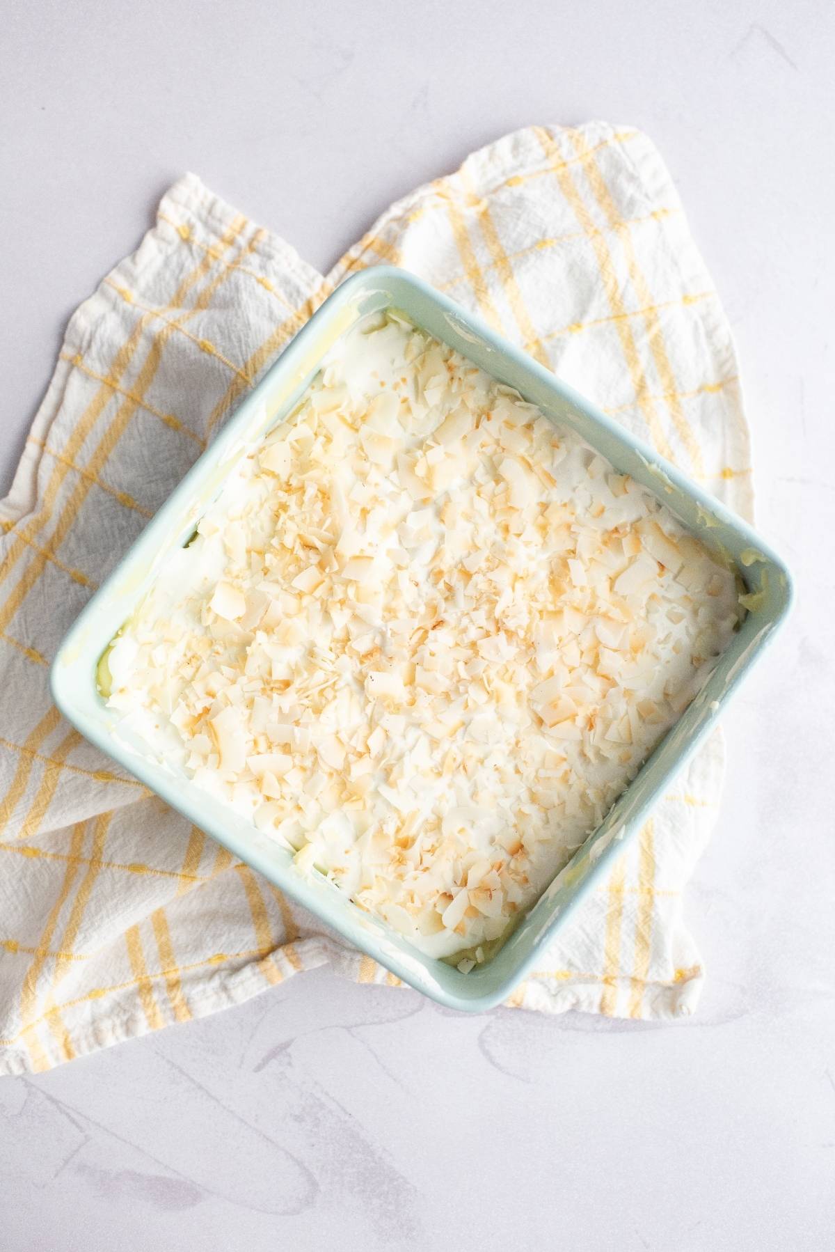 A square dish of pineapple lush, topped with shredded coconut, sits on a yellow and white checkered cloth—perfect as a creamy summer dessert for a BBQ.