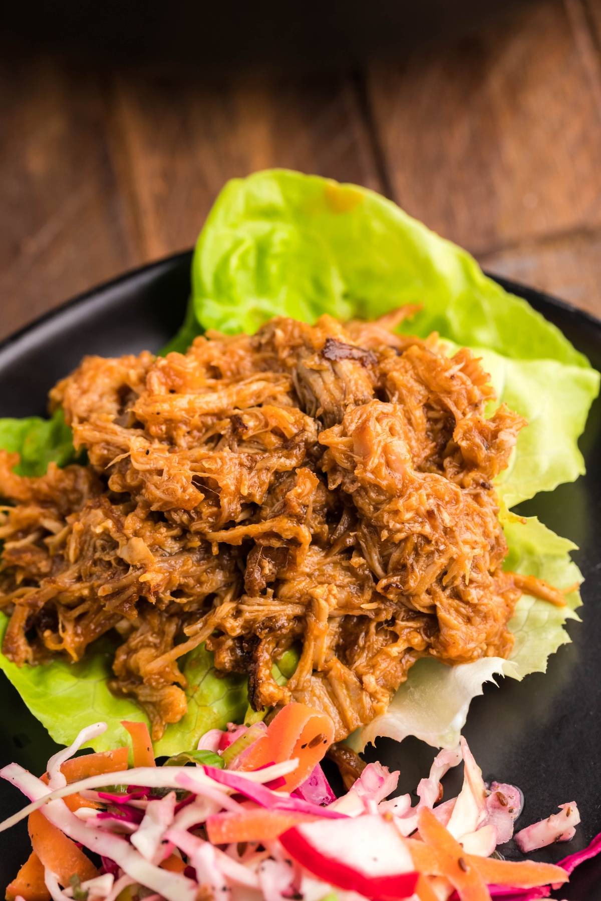 Slow cooked pork shoulder shredded for pulled pork in lettuce wraps, served with a side of colorful coleslaw on a black plate.