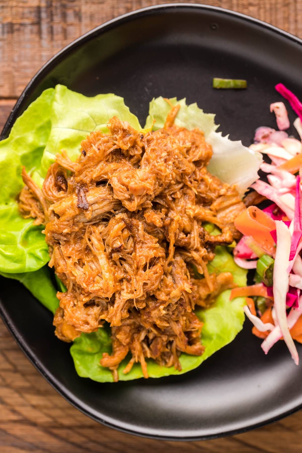 Pulled pork in a Dutch oven served on lettuce leaves with a side of colorful Dairy Free Vegan Coleslaw, all beautifully arranged on a black plate.