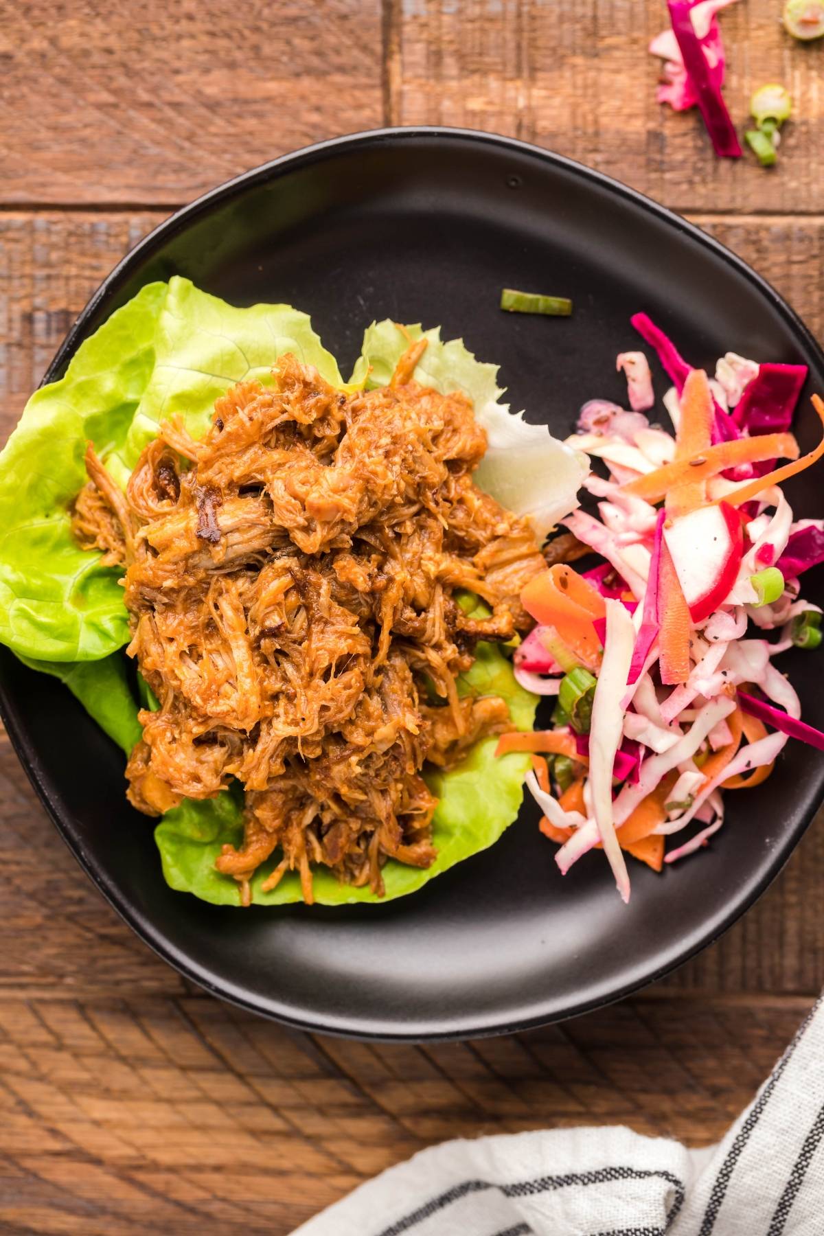 Pulled pork in a Dutch oven on lettuce leaves, topped with colorful dairy free coleslaw, served on a black plate atop a wooden table.