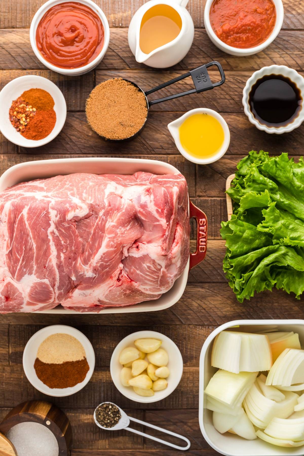 Raw pork, seasonings, sauces, garlic, onions, and leafy lettuce are arranged on a wooden table—perfect for making pulled pork in lettuce wraps.