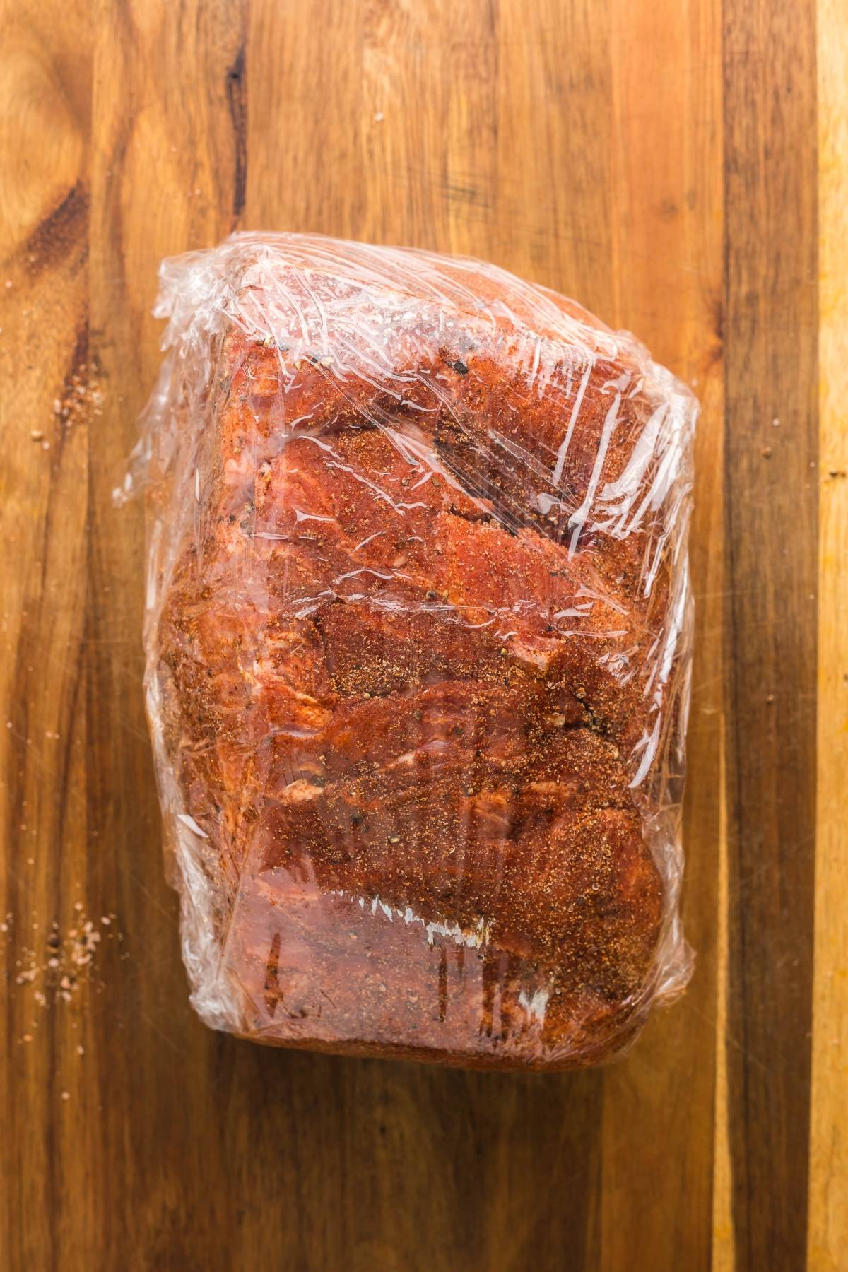 A seasoned piece of meat wrapped in plastic wrap on a wooden cutting board—perfect for making pulled pork in a Dutch oven.