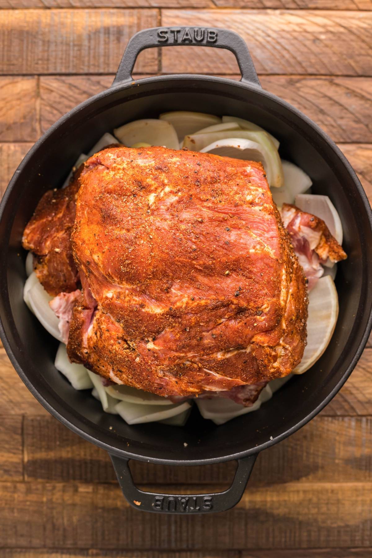 Seasoned pork roast sits atop sliced onions in a black Staub pot, ready to become tender pulled pork in a dutch oven—perfect when paired with dairy free vegan coleslaw.