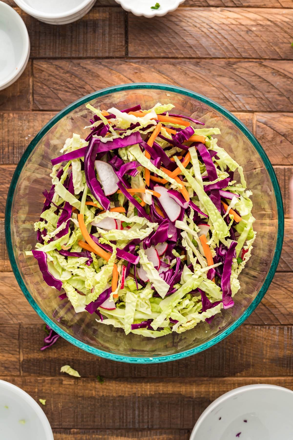 A glass bowl filled with Dairy Free Vegan Coleslaw—shredded green and purple cabbage, carrots, and radishes—sits on a wooden table.