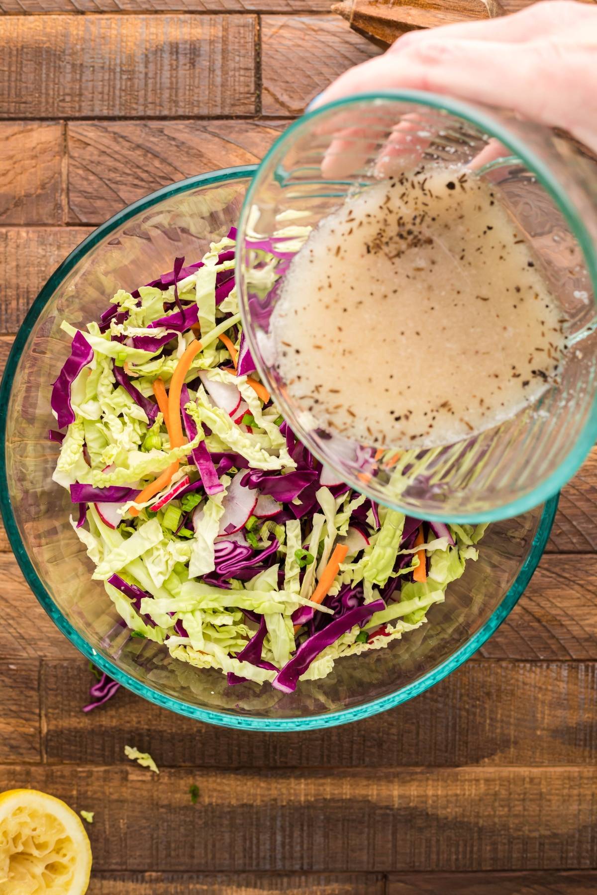 A hand pours dressing over a bowl of shredded cabbage and carrots on a wooden surface, the perfect start for Pulled Pork and Dairy Free Coleslaw.