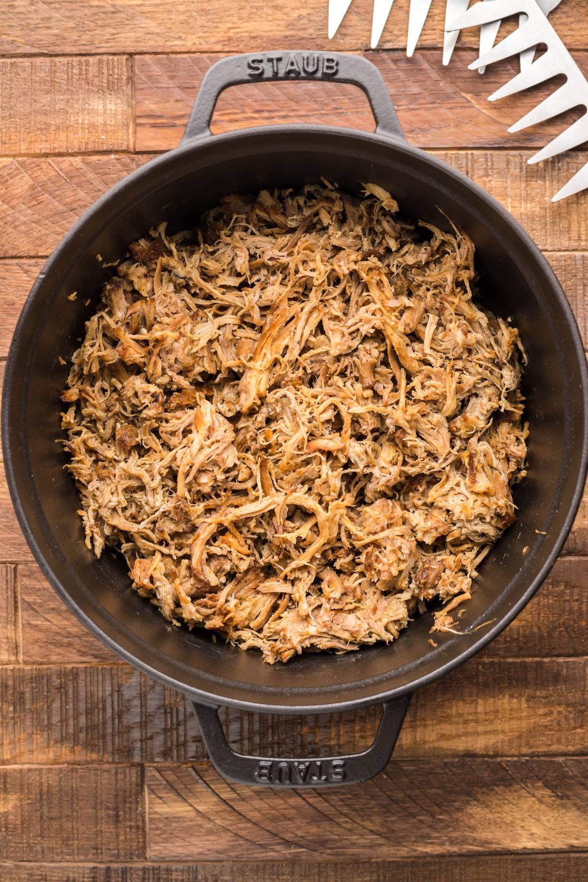 Shredded slow cooked pork shoulder in a black pot on a wooden surface, with a white meat shredder beside it—perfect for pairing with Dairy Free Vegan Coleslaw.