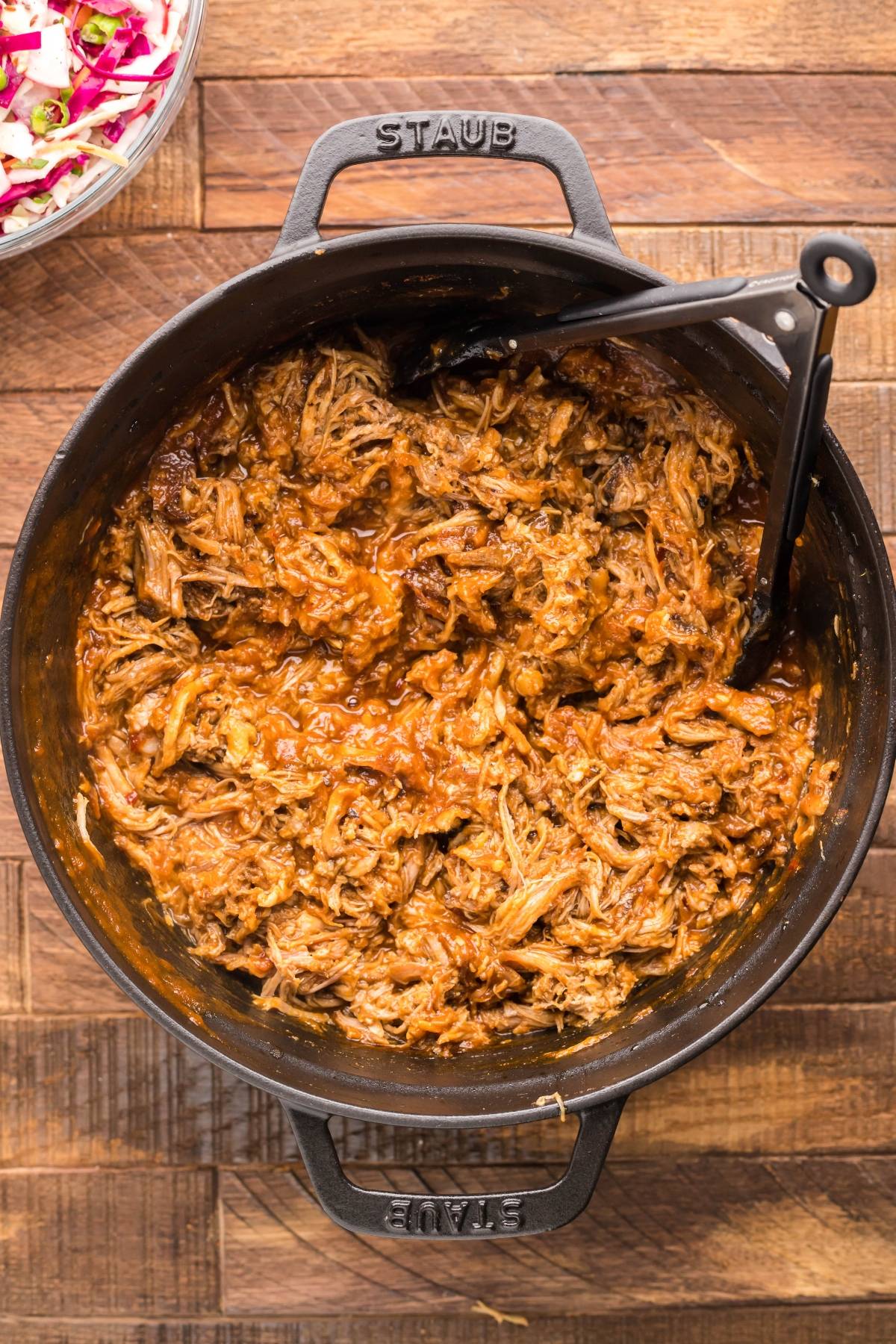 Slow cooked pork shoulder is shredded for barbecue pulled pork in a Dutch oven, served hot with a spoon on a wooden table.