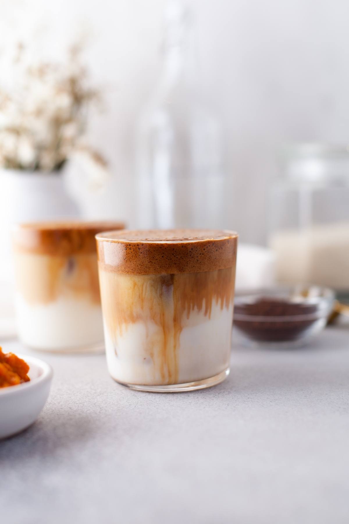 Glass of pumpkin whipped coffee with milk, featuring a creamy blend of pumpkin puree and instant coffee; blurred background with another glass and cozy kitchen items captures the essence of autumn coffee flavour.