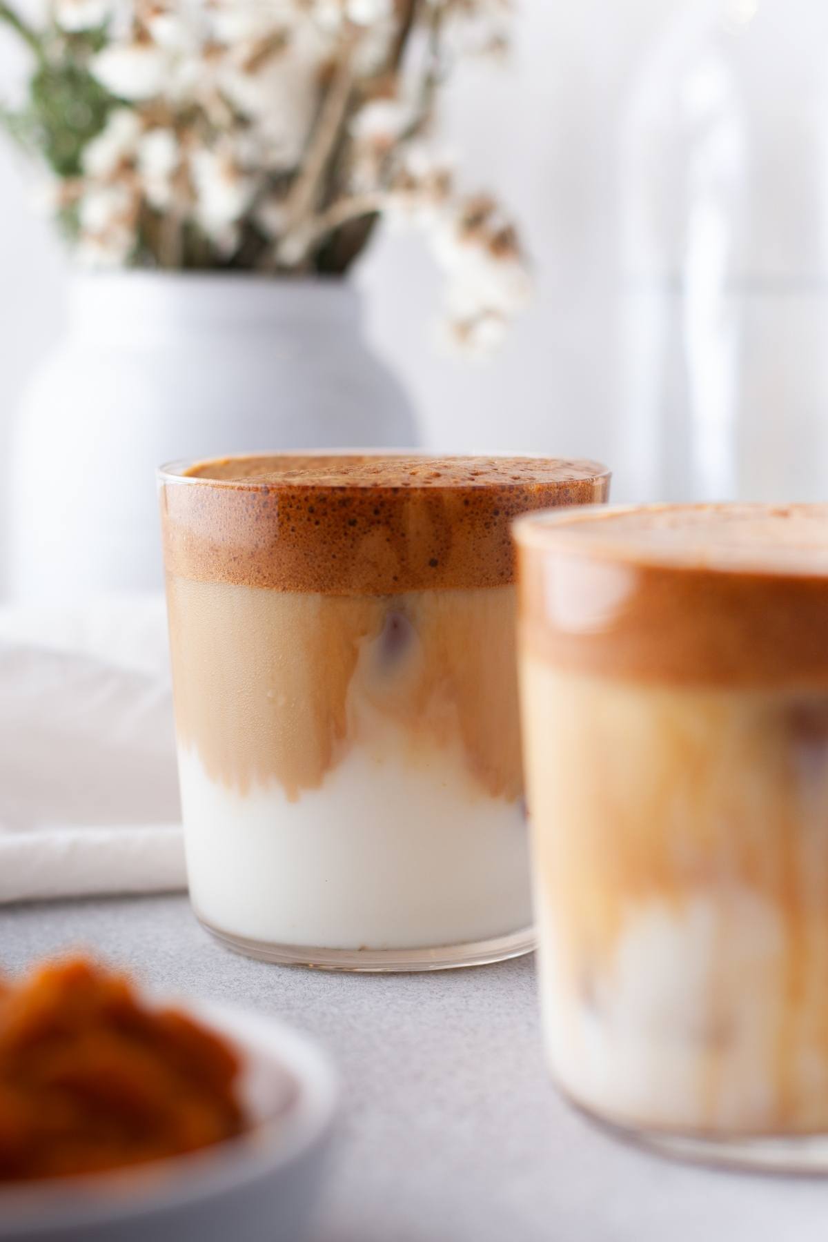 Two glasses of iced coffee with creamy milk layers, featuring a touch of autumn coffee flavour—perfect for trying a seasonal coffee recipe. Flowers and a white vase add charm in the background.