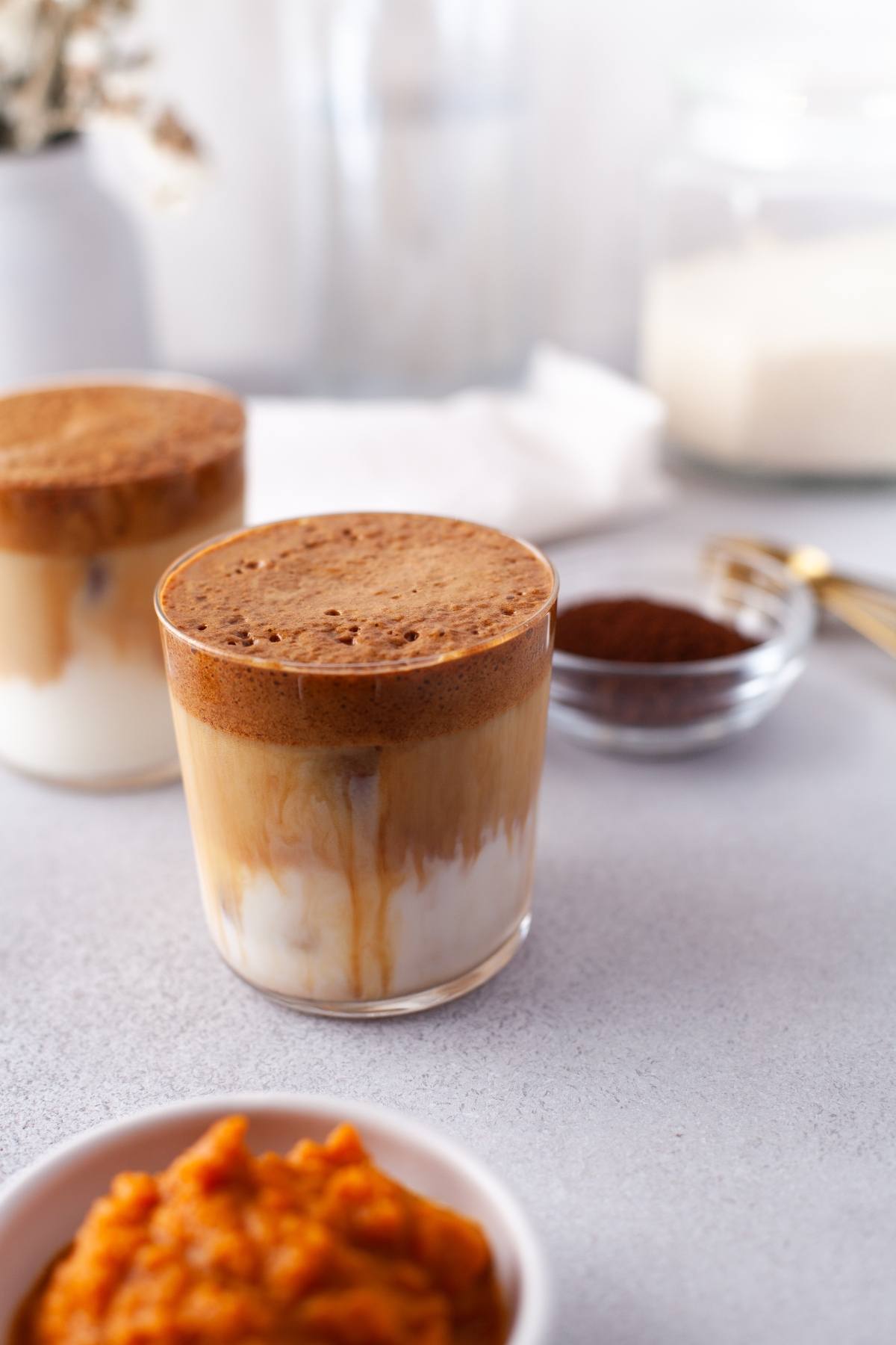 Glass of whipped coffee on milk with pumpkin puree and instant coffee in the foreground, perfect for an autumn coffee flavour; coffee grounds rest in the background on a light surface.