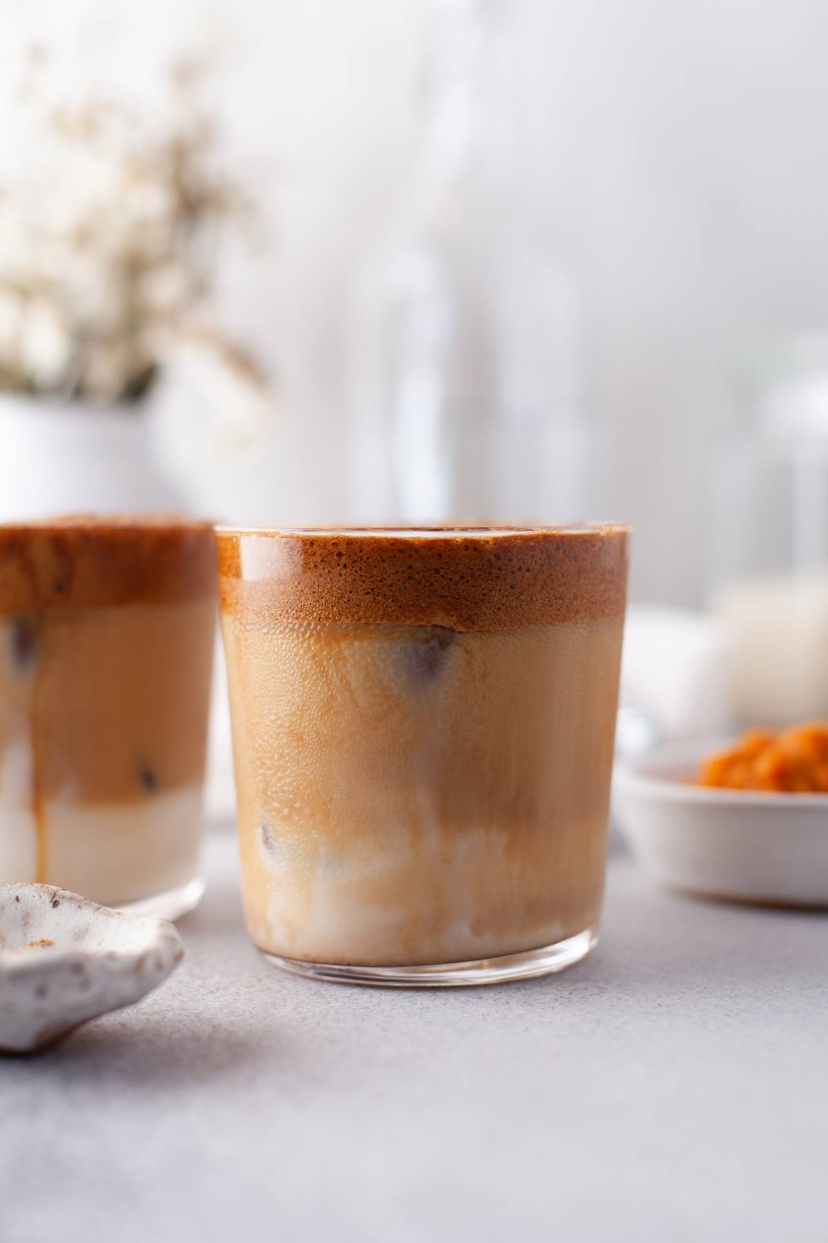 A glass of iced Pumpkin Spice Whipped Dalgona Coffee with a creamy, frothy layer on top, set on a light-colored surface—a delightful seasonal coffee recipe perfect for fall.