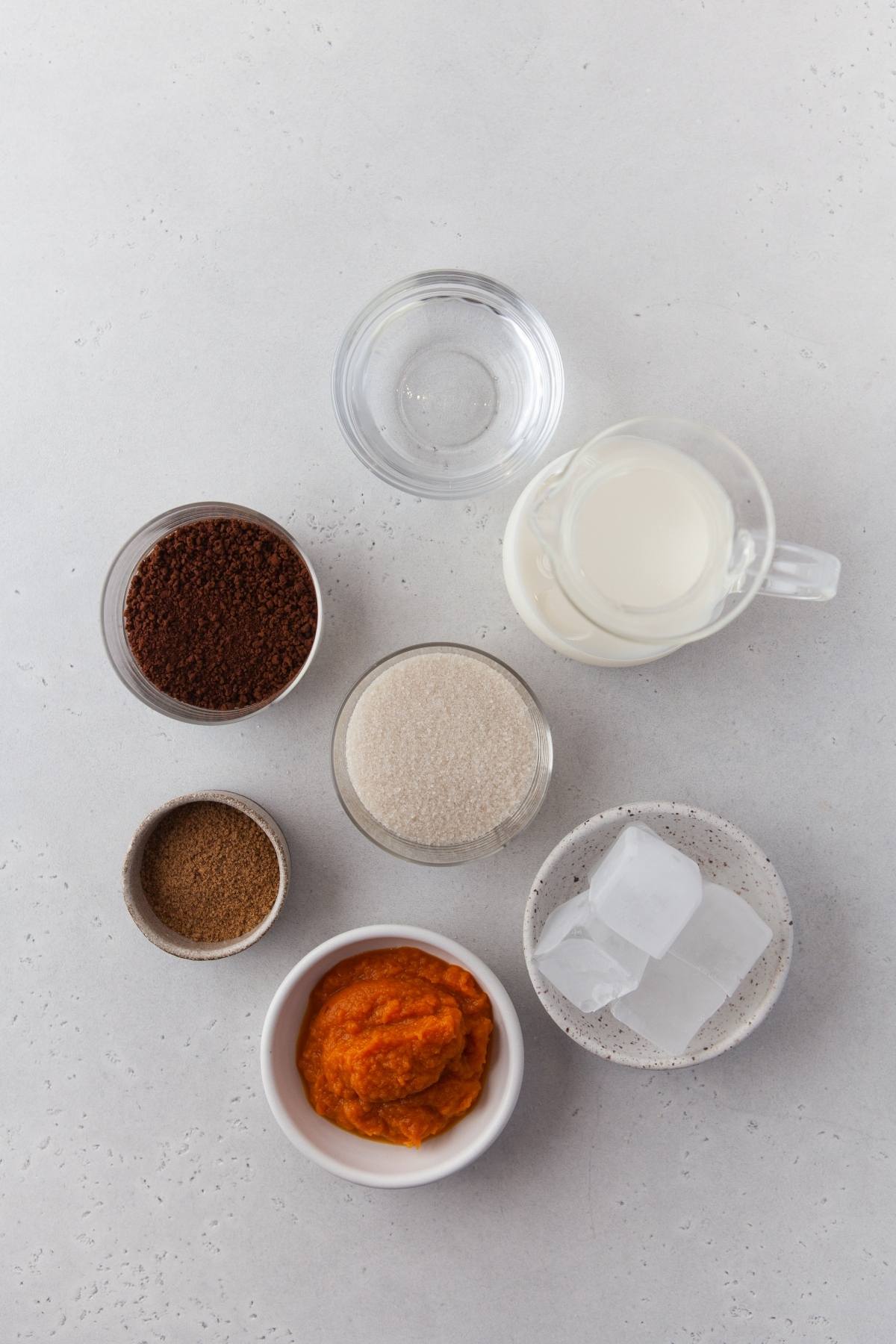 Top-down view of bowls with pumpkin puree and instant coffee, sugar, spices, milk, water, and ice on a light surface—perfect for crafting a pumpkin whipped coffee or your favorite seasonal coffee recipe.