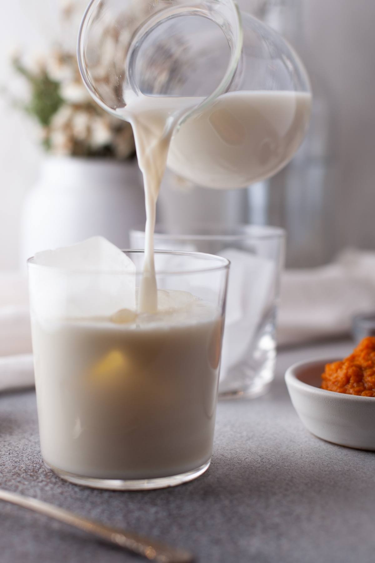 Milk being poured from a glass pitcher into a glass with ice, perfect for serving your favorite autumn coffee flavour or seasonal coffee recipe, with a blurred background and a bowl nearby.