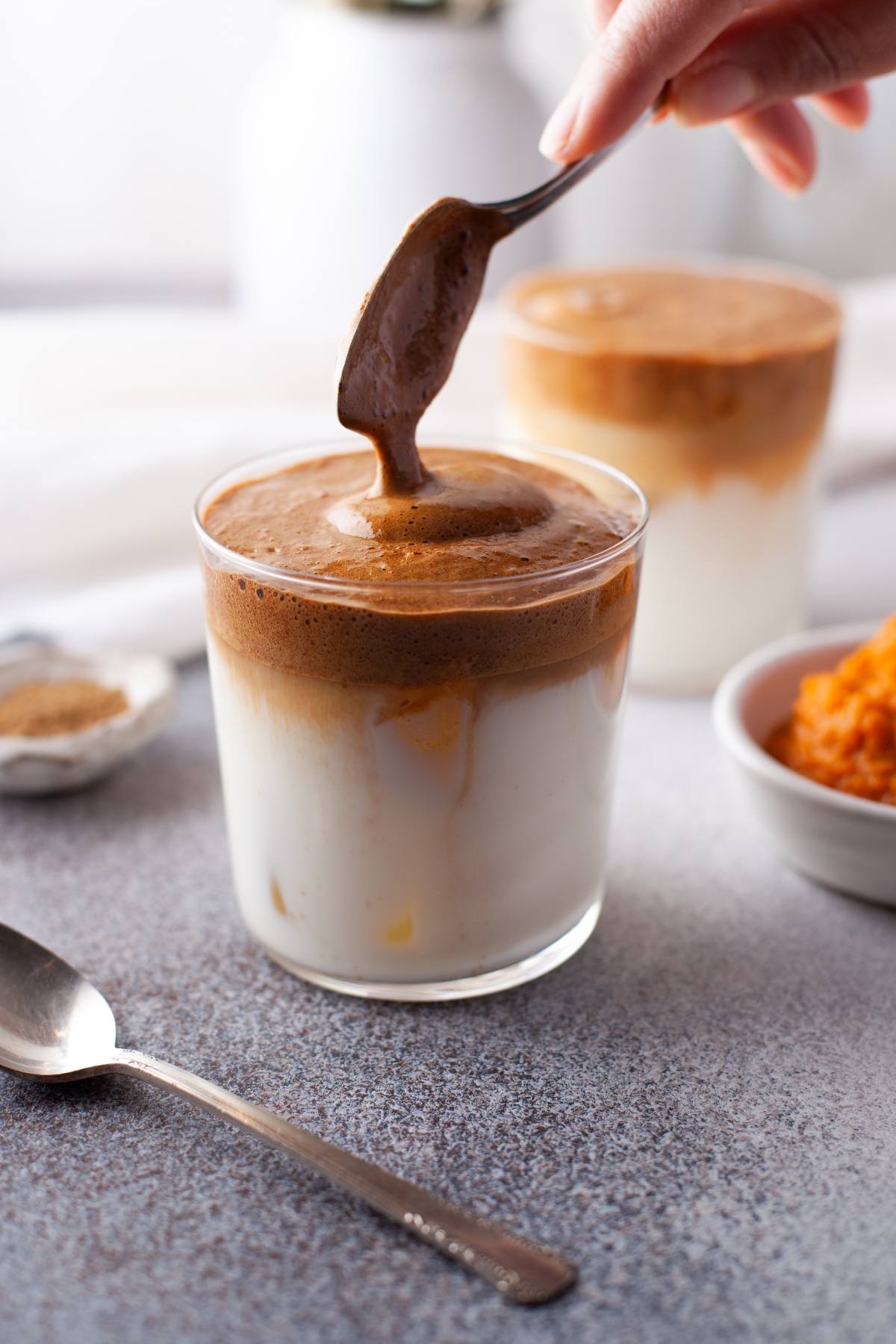 A hand adds Pumpkin Spice Whipped Dalgona Coffee to a glass of milk, with a spoon and bowl nearby on a gray surface—an irresistible seasonal coffee recipe perfect for autumn.