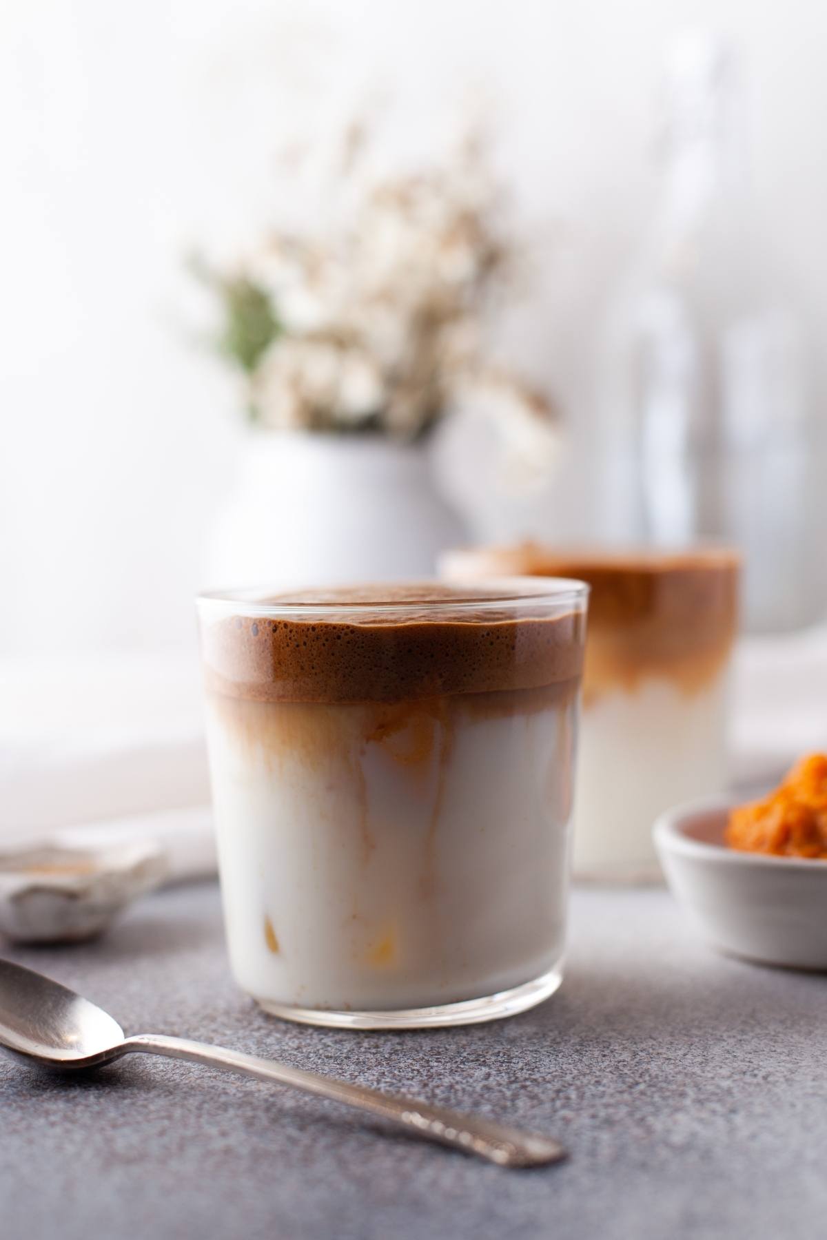 A glass of whipped coffee with milk sits on a table, next to a spoon and small bowl, with blurred flowers behind—an inviting seasonal coffee recipe perfect for autumn.