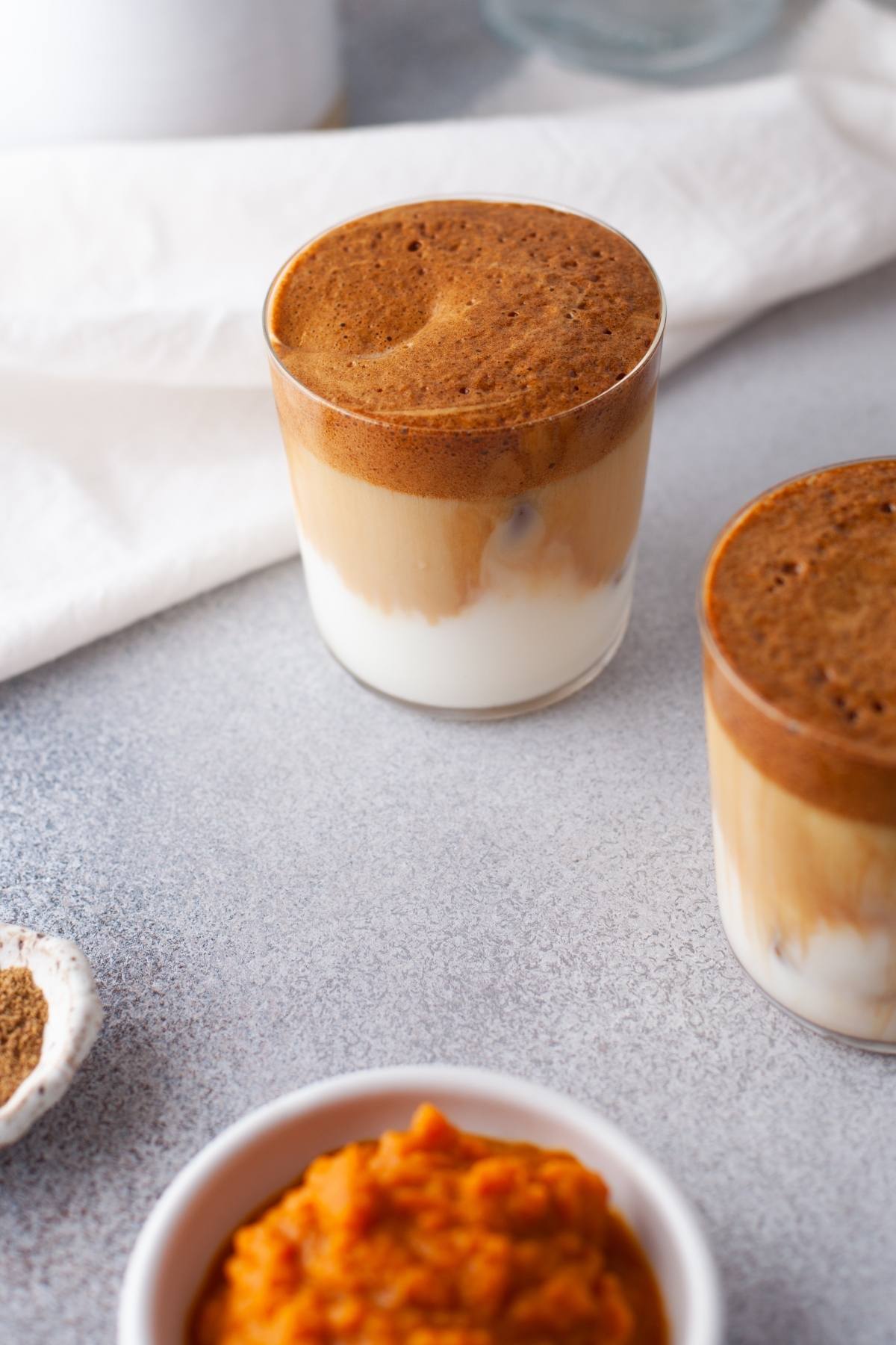 Two glasses of Pumpkin Spice Whipped Dalgona Coffee sit on a gray surface, with a bowl of pumpkin puree and instant coffee in the foreground—a perfect seasonal coffee recipe.