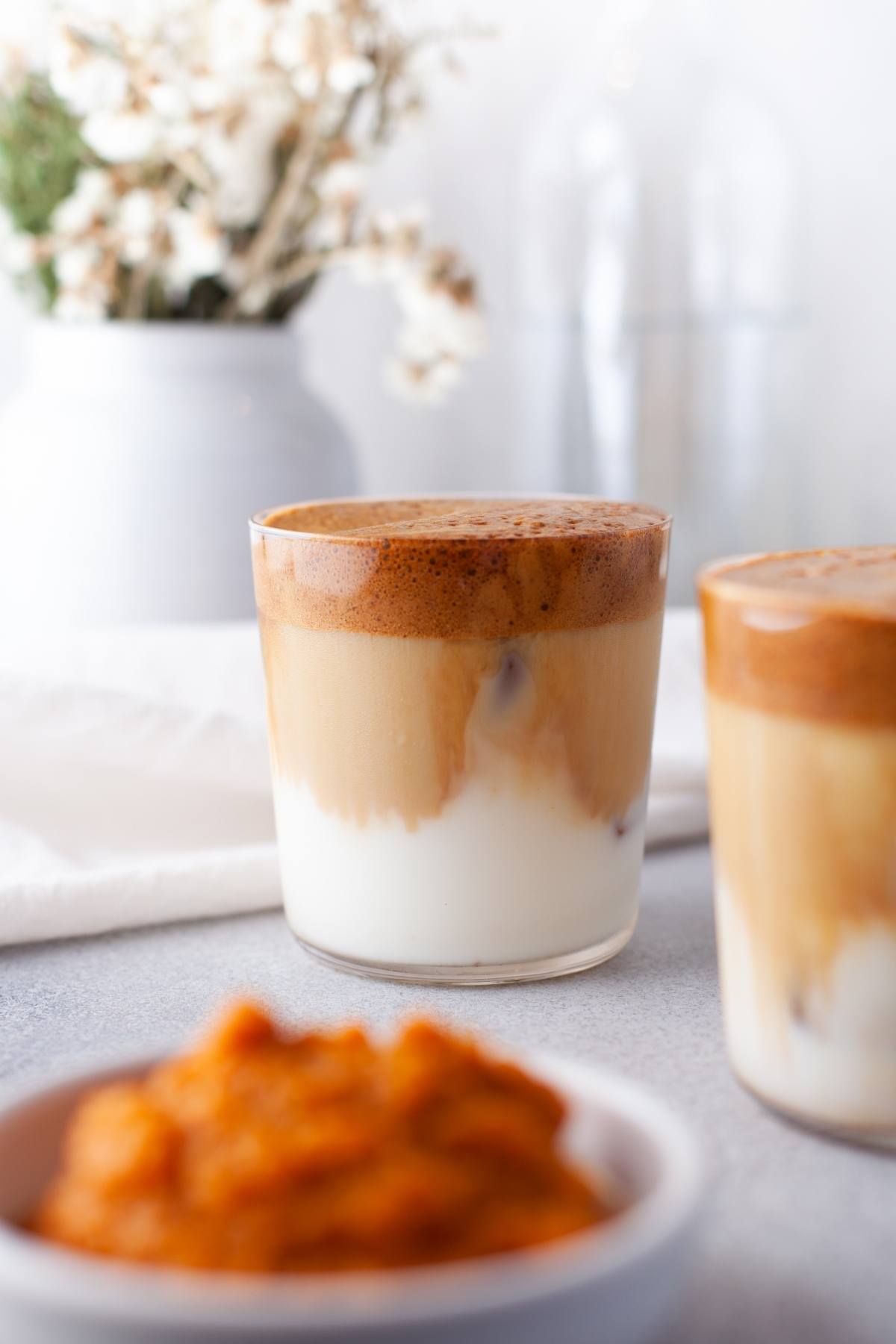 A glass of Pumpkin Spice Whipped Dalgona Coffee with milk, with a blurred bowl of pumpkin puree and flowers in the background, captures the essence of autumn coffee flavour.