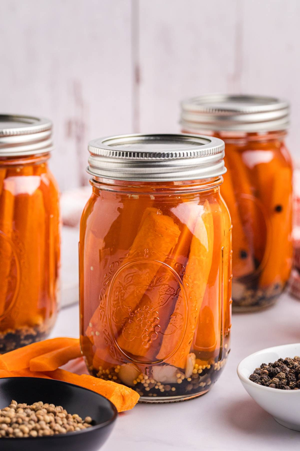 Three glass jars filled with pickled carrots and spices, with lids sealed, on a white surface.