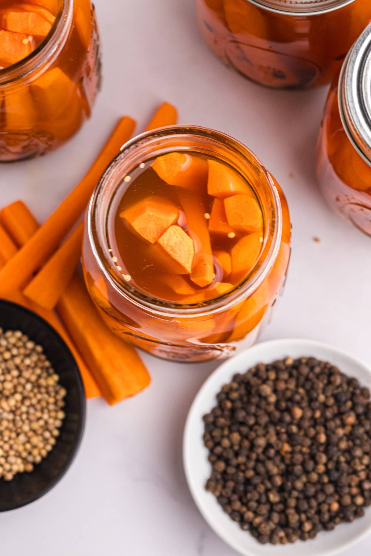 Open jar of pickled carrots with spices, surrounded by carrot sticks and bowls of peppercorns and seeds.