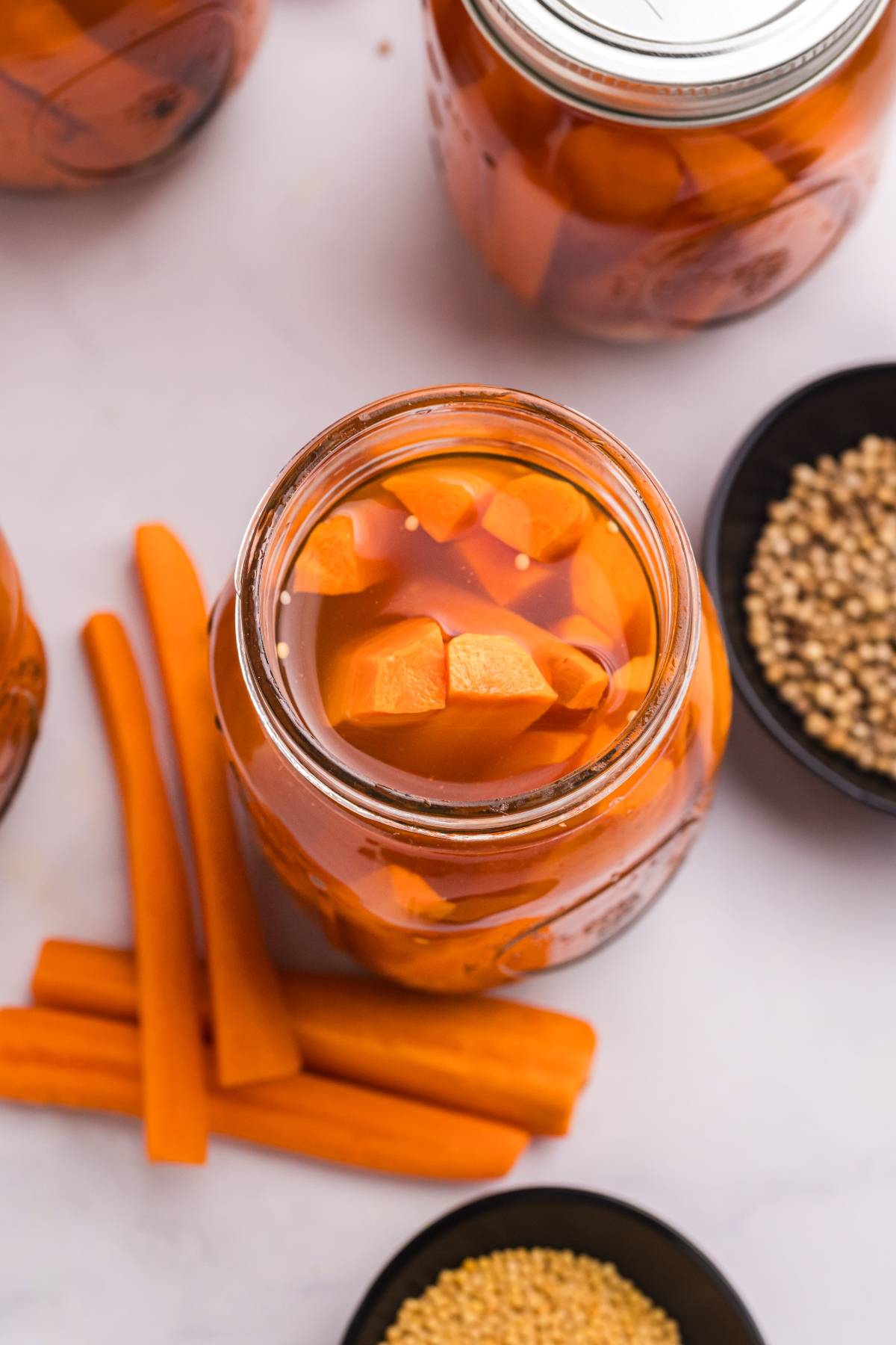Open jar of pickled carrots with carrot sticks and bowls of seeds on a white surface.