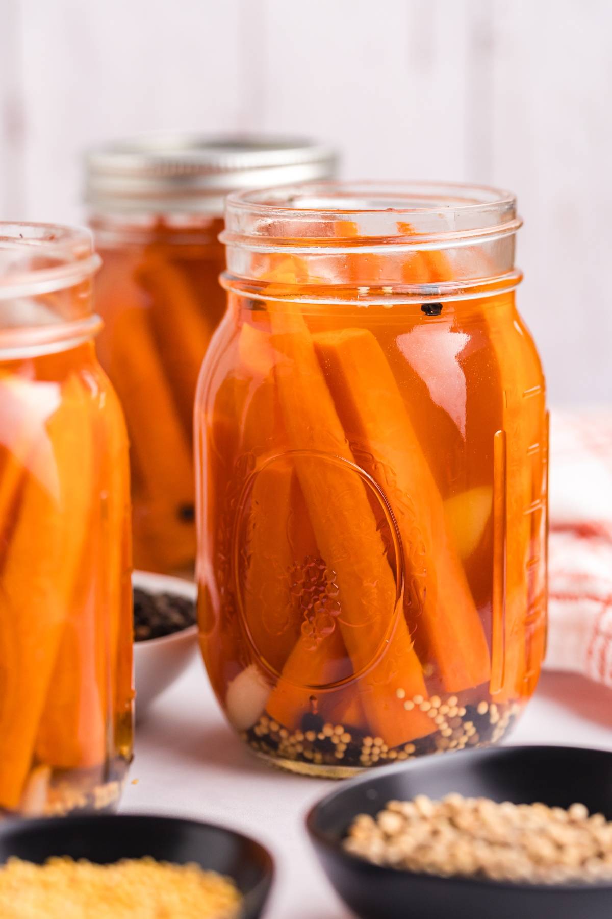 Three jars filled with pickled carrot sticks and spices, with seasoning bowls in the foreground.