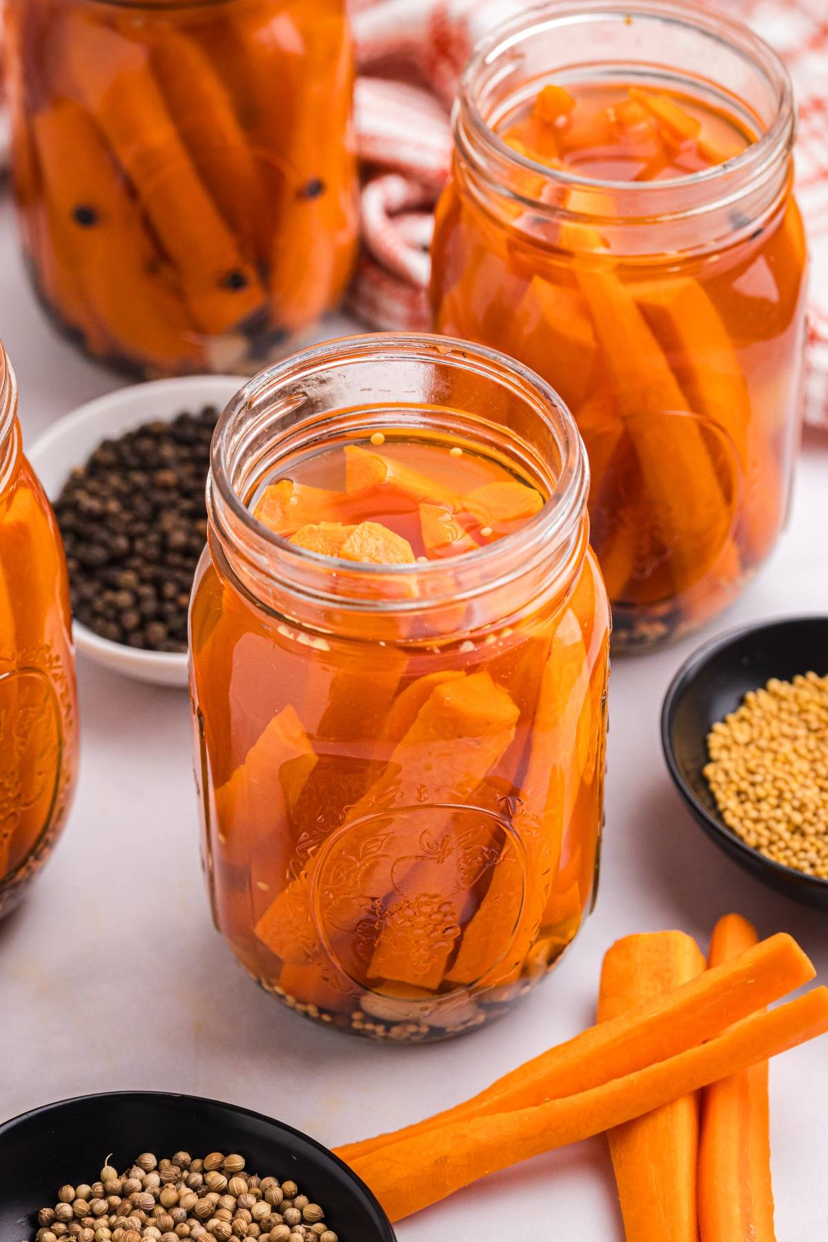Jars filled with pickled carrot sticks, surrounded by spices and fresh carrot pieces on a white surface.