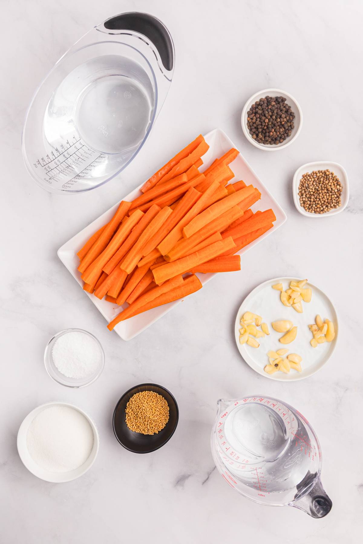 Carrot sticks, garlic, salt, sugar, peppercorns, coriander, mustard seeds, and water on a white surface.