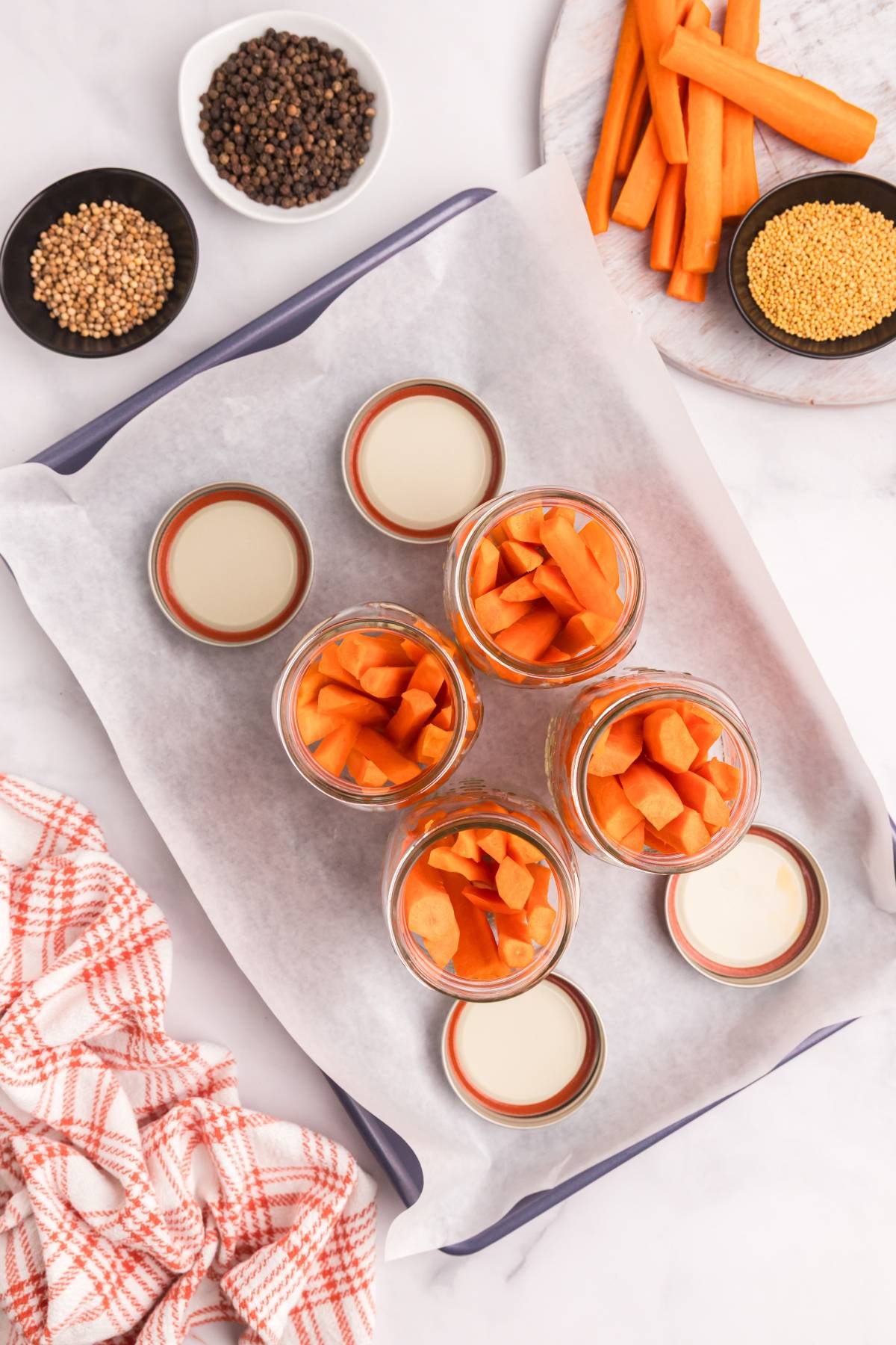 Jars filled with carrot sticks on a tray, surrounded by spices, carrot pieces, and jar lids.
