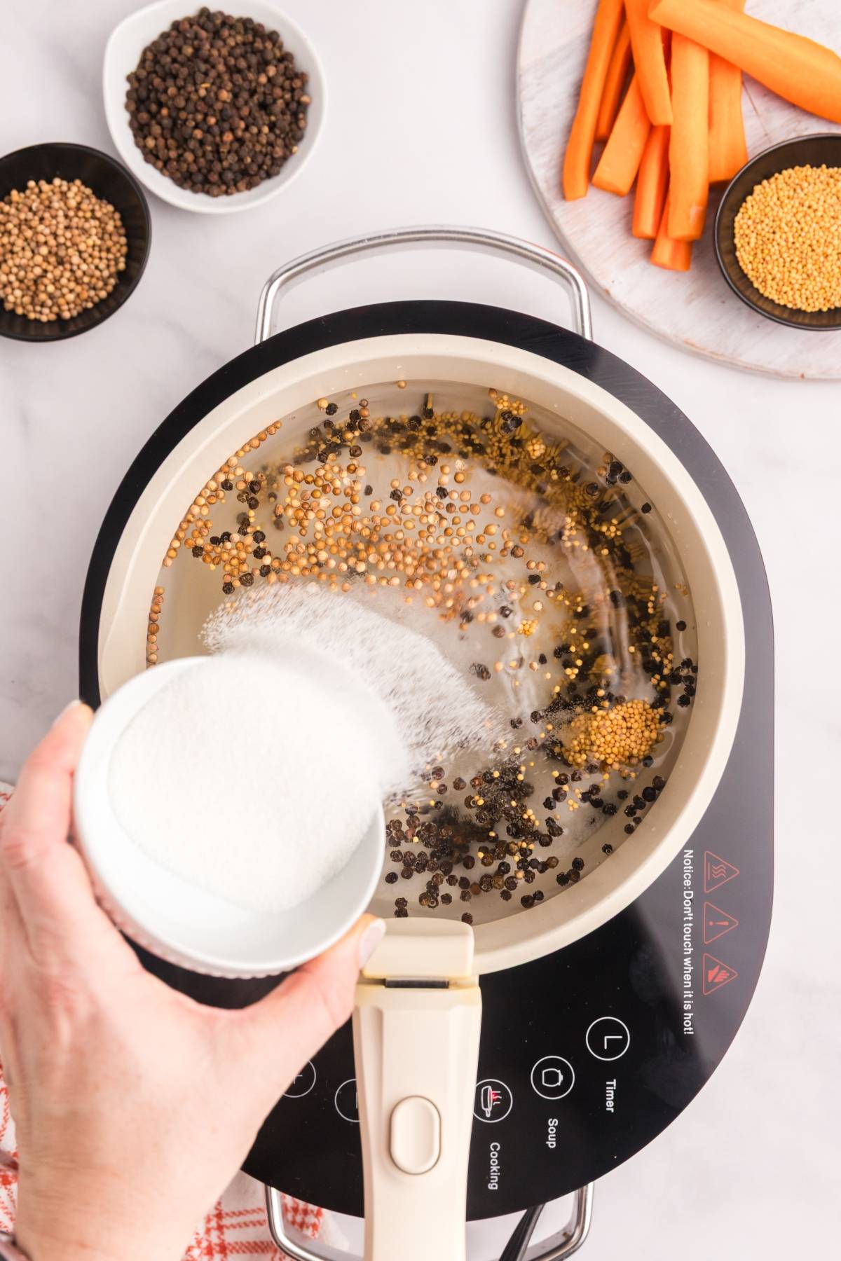 A hand pours sugar into a saucepan with peppercorns and mustard seeds on a stovetop, with carrots nearby.