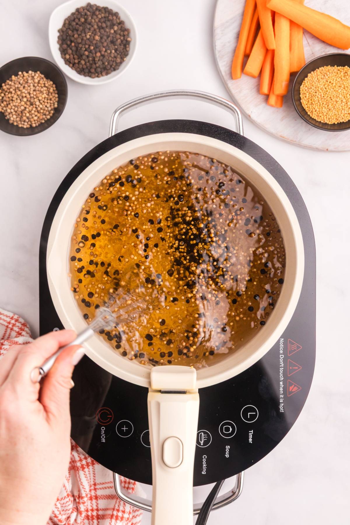 A hand stirs pickling brine with peppercorns and seeds in a pot; carrots and seeds are nearby.