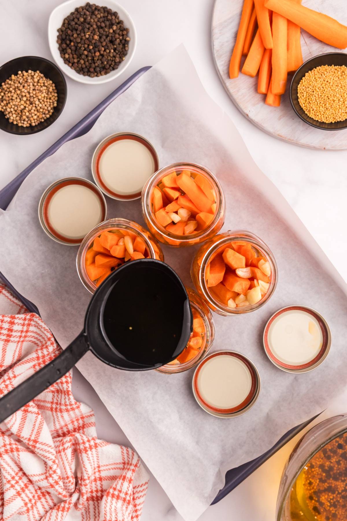 A ladle pours liquid into jars filled with chopped carrots and garlic, surrounded by spices and lids.