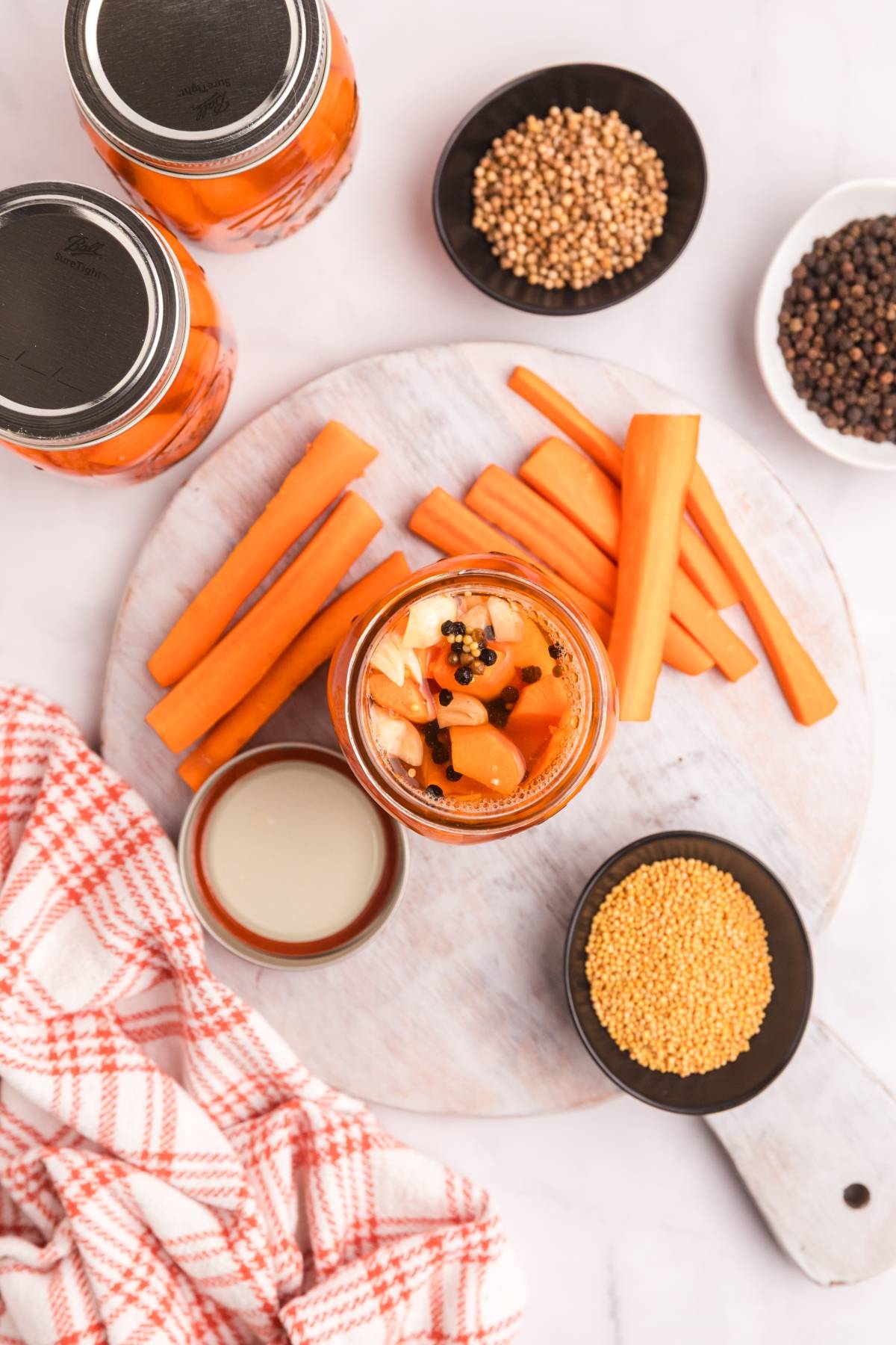 Top view of sliced carrots, a jar of pickled carrots, spices, and a red plaid towel on a white board.