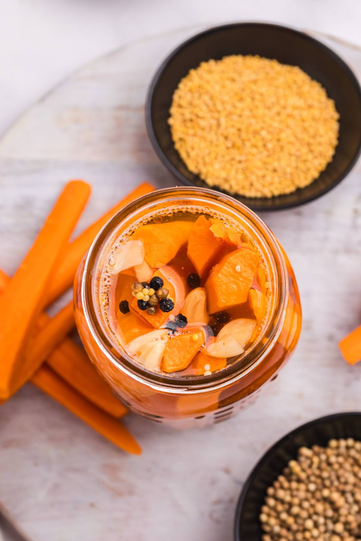 A jar of pickled carrots with garlic and spices, surrounded by carrot sticks and bowls of seeds.