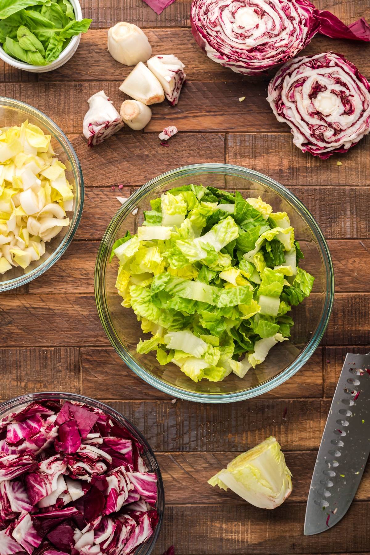 Chopped lettuce and radicchio in bowls on a wooden table with a knife and fresh vegetables nearby—perfect for making a simple radicchio salad.