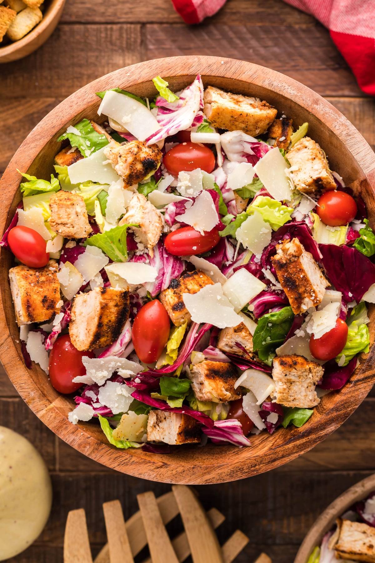 Wooden bowl with summer BBQ chicken salad, cherry tomatoes, mixed greens, and shaved parmesan cheese on a wooden table.