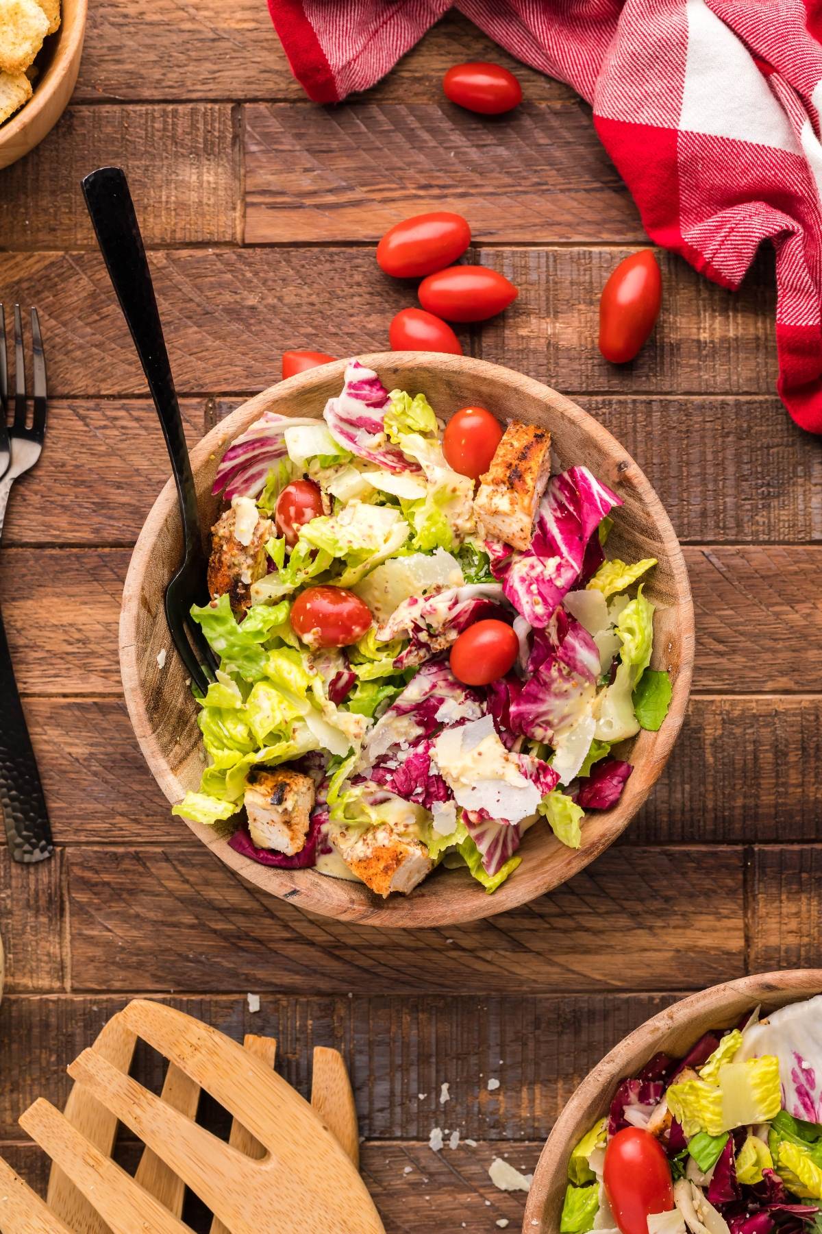 A wooden bowl of summer BBQ chicken salad with lettuce, grape tomatoes, croutons, and a fork on a wooden table.